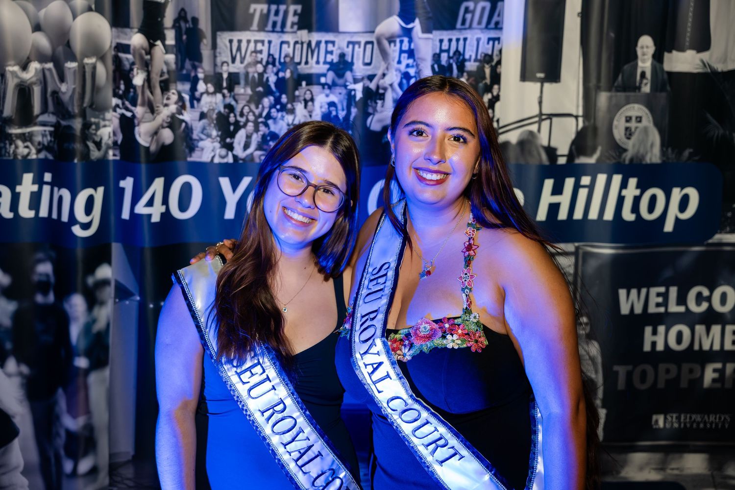 Beauty pageant contestants wearing blue sashes pose together at a nighttime event with dark lighting.