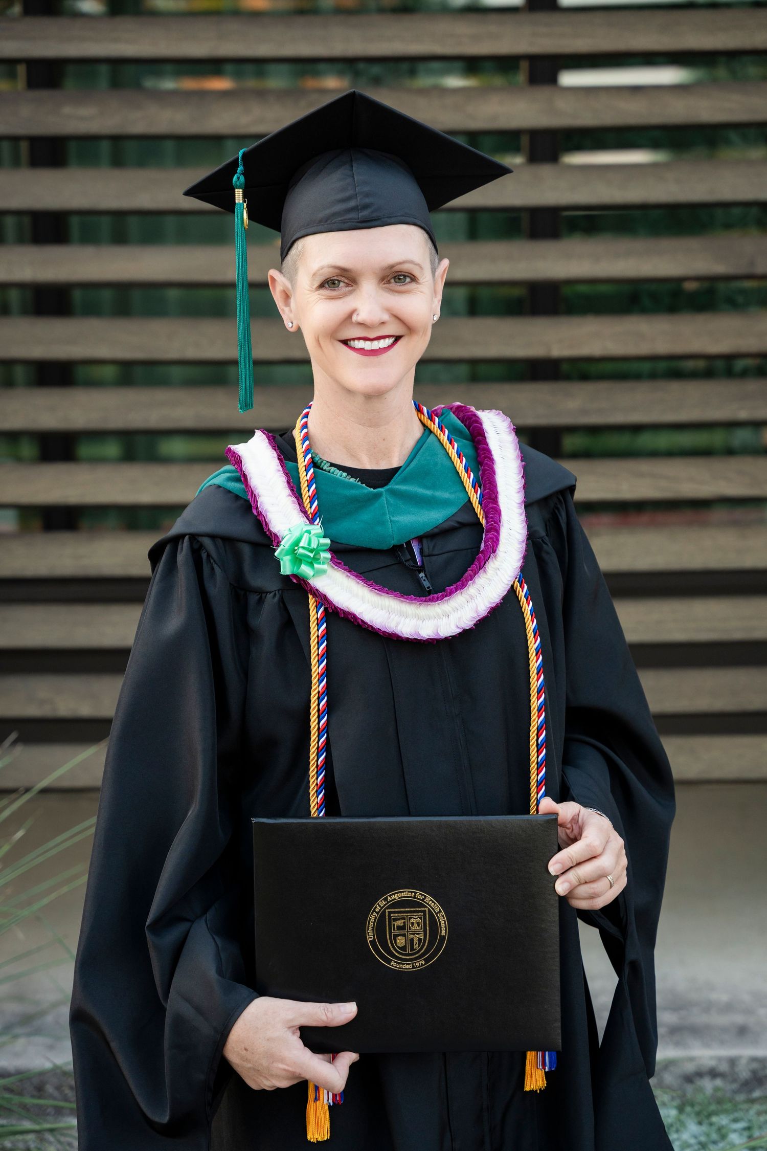 Graduate in black cap and gown holding diploma with green tassel and colorful graduation stole at commencement ceremony.