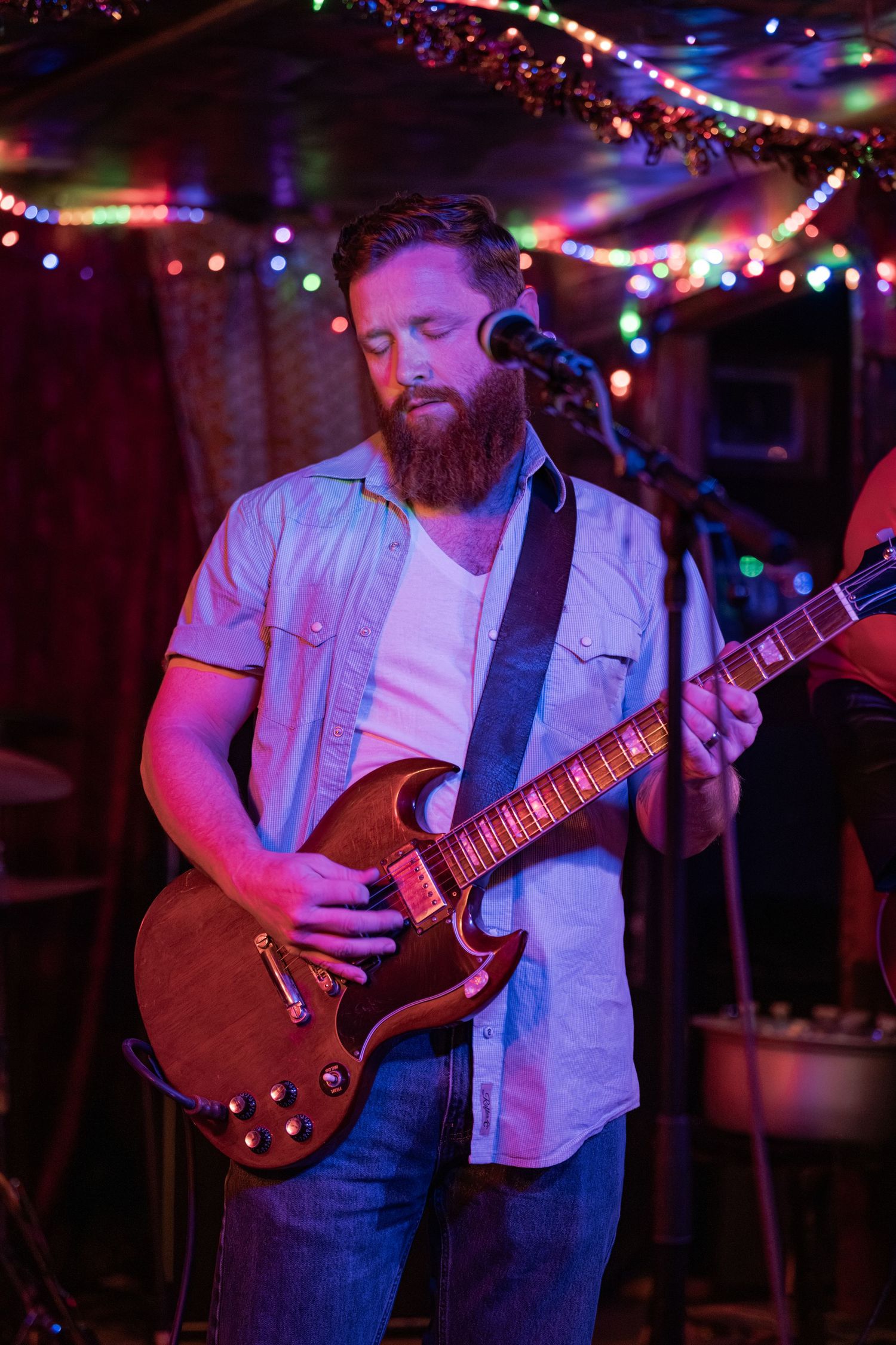 Musician in denim overalls plays electric guitar under colorful string lights at a dimly lit bar venue.