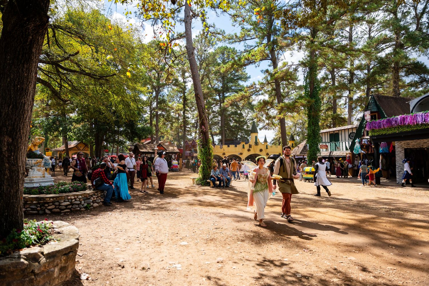 Visitors walk along a shaded dirt path at a Renaissance festival with vendor booths and trees lining the way.