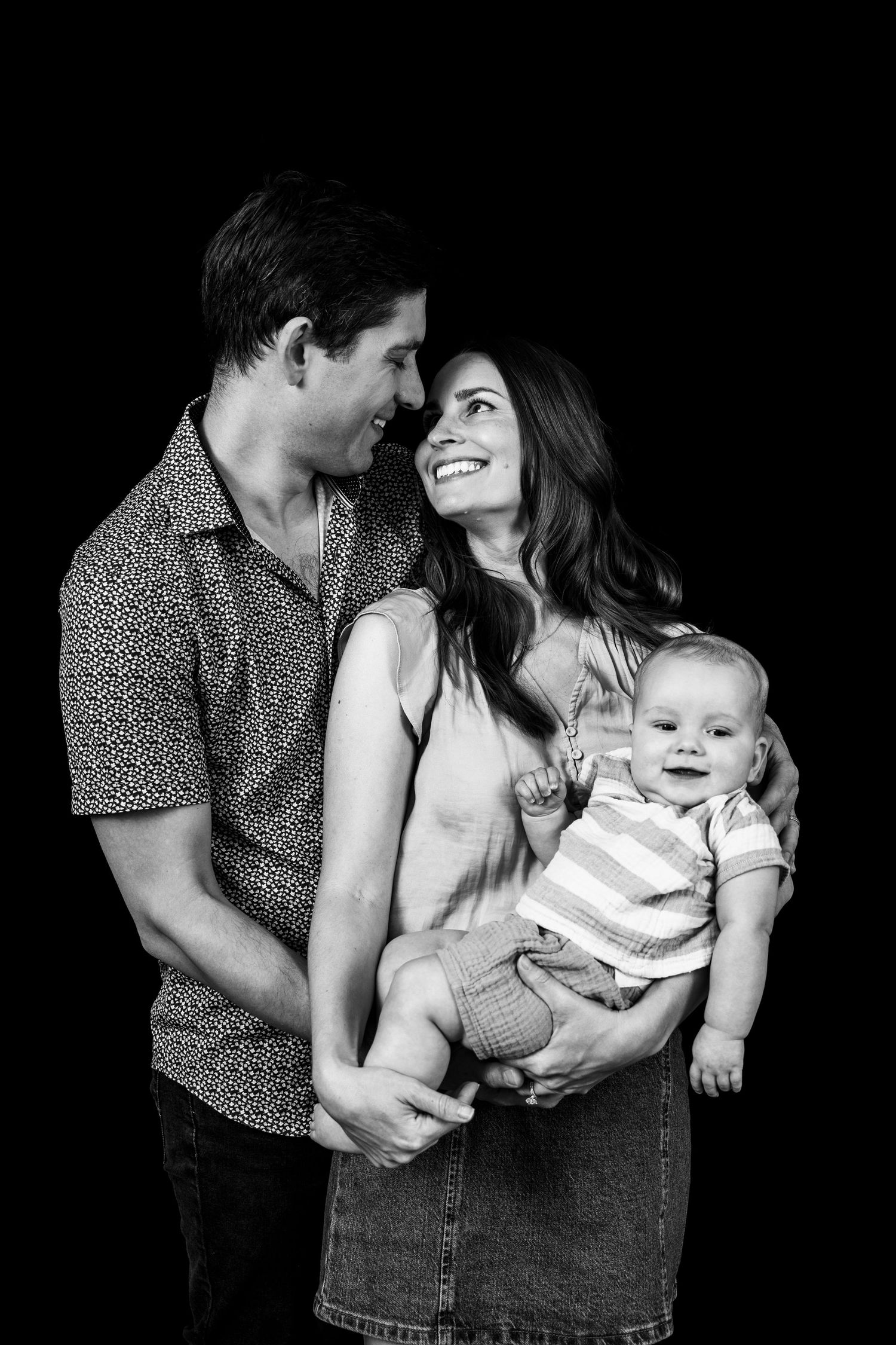 Black and white portrait of a couple sharing a loving moment while holding their smiling baby against a dark background.