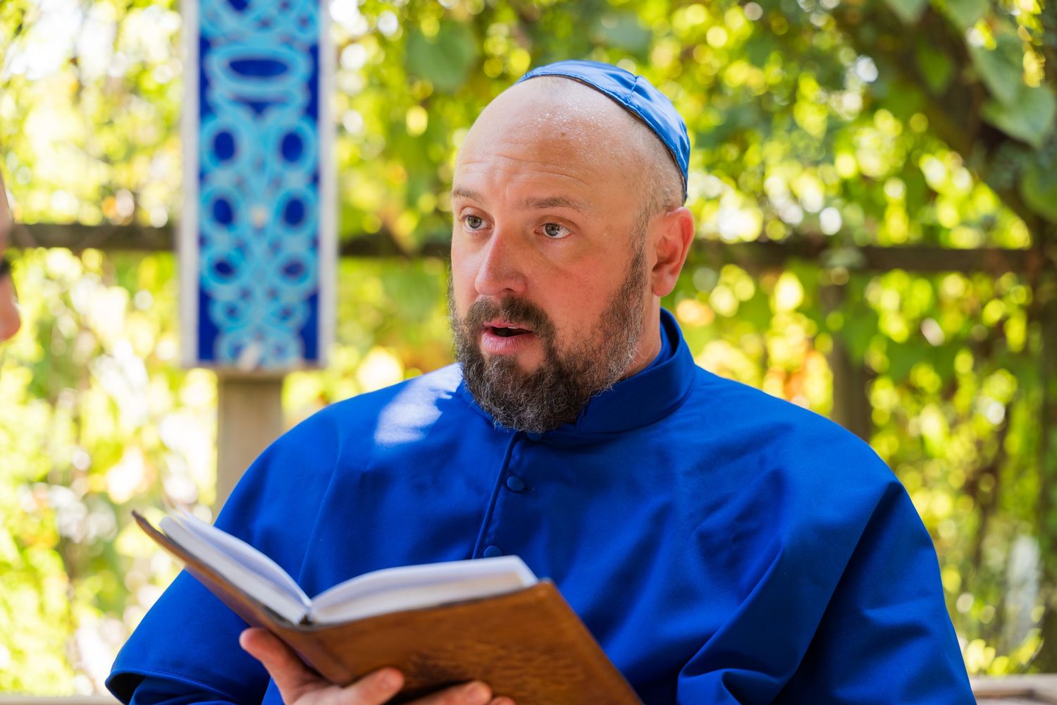 A person in a bright blue shirt reads from a book while standing outdoors with green foliage in the background.