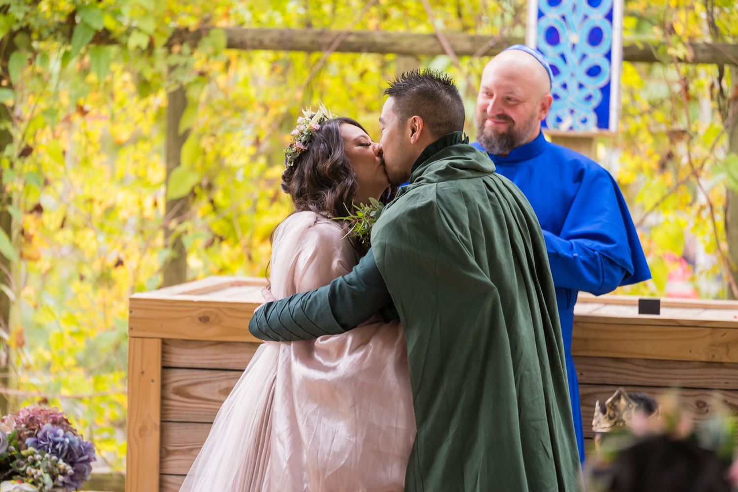 A couple embraces outdoors during autumn with yellow foliage in the background and wooden railings nearby.