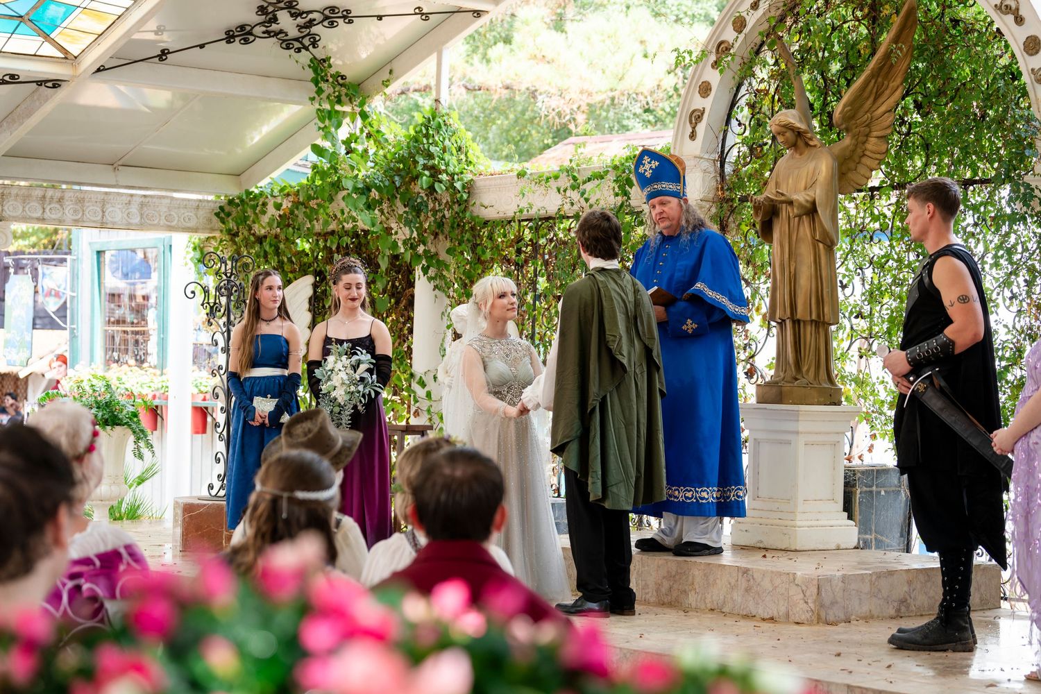 Wedding guests gather under an outdoor pavilion decorated with greenery and flowers during a summer celebration.
