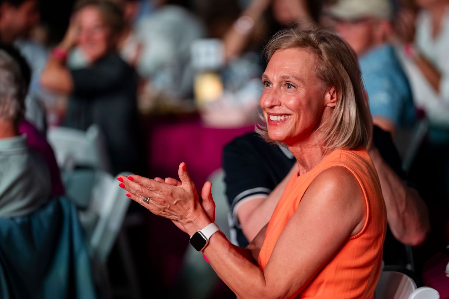 A person in an orange shirt laughs and claps while sitting in a crowded audience.
