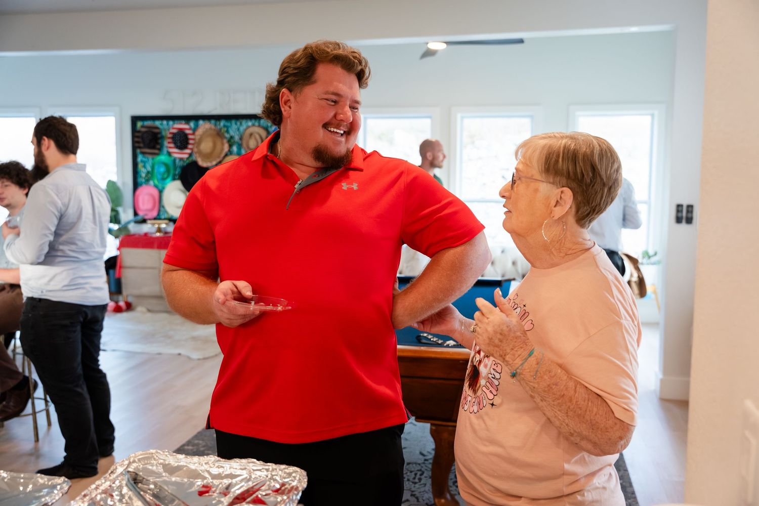 A man in a red polo shirt laughs with an elderly woman at a casual indoor gathering or party.