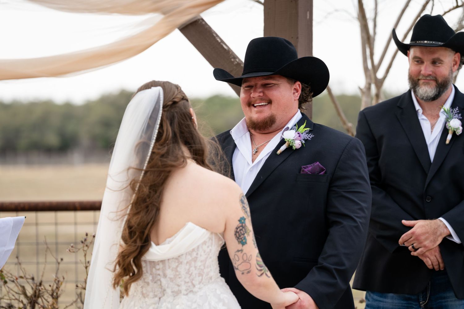 Groom in black cowboy hat smiles at bride during outdoor country wedding ceremony with rustic wooden arch.