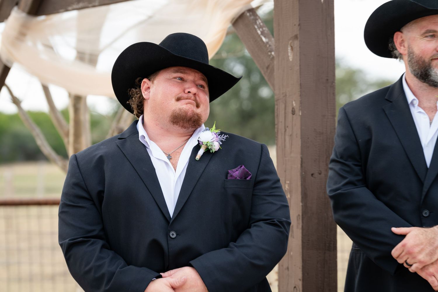 Man in black suit and cowboy hat with boutonniere at outdoor western wedding ceremony.