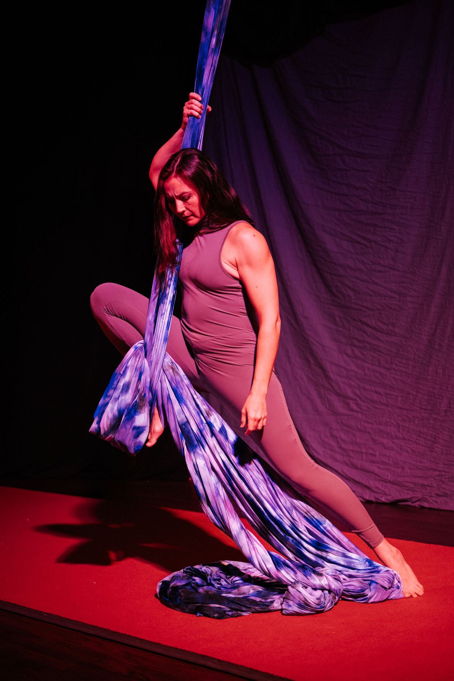 Aerial silk dancer in gray bodysuit performing on red stage with blue tie-dye fabric under dramatic purple lighting.