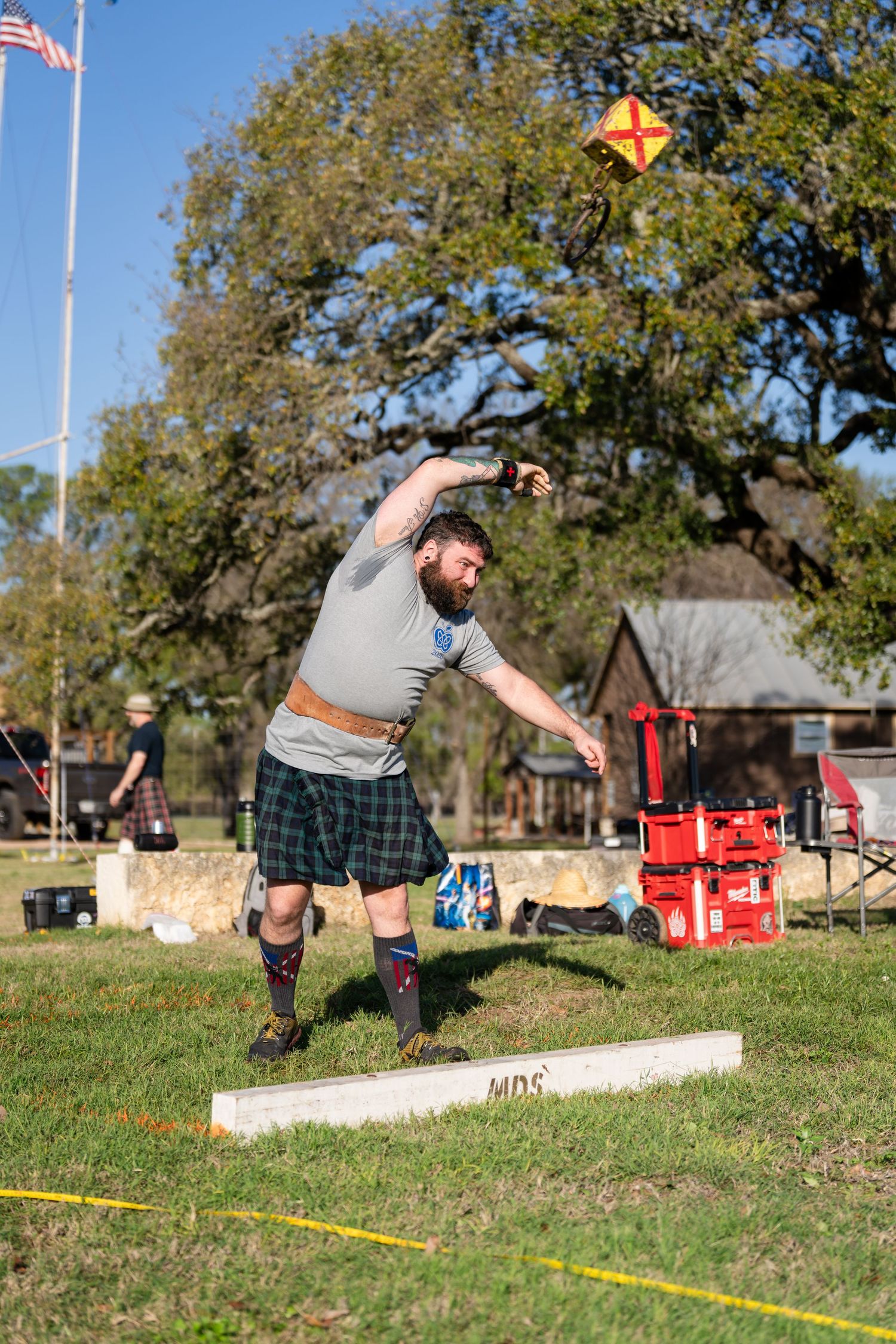 Bearded man in tartan kilt competing in Highland Games, throwing object overhead on grassy field with trees and flag in background.