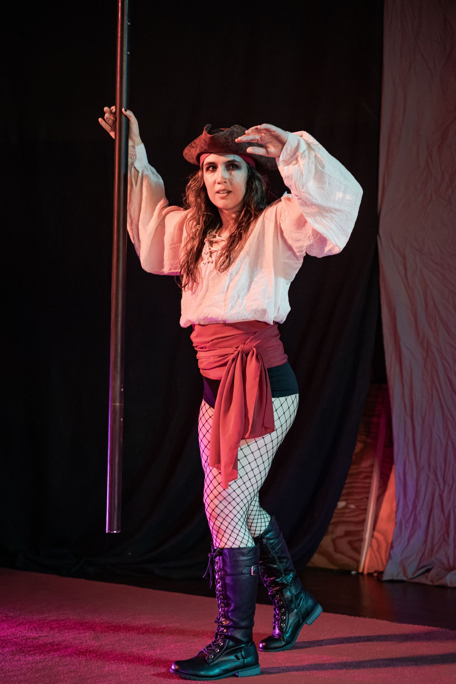 Woman in pirate costume with white blouse, red sash, fishnet tights, and boots poses by a pole on stage.