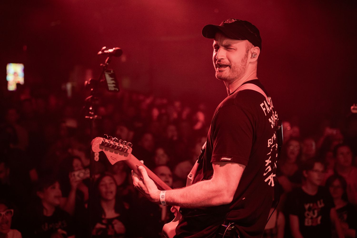 Guitarist performing on stage under red lights, facing a crowd at a live concert venue.