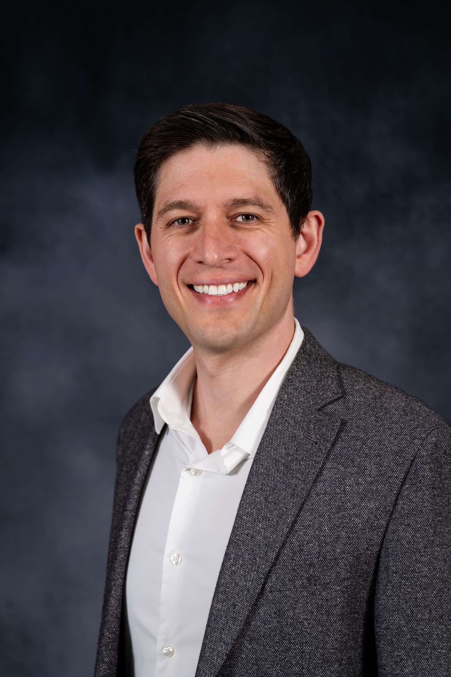 Professional headshot of a smiling young man in a gray blazer and white dress shirt against a dark backdrop.