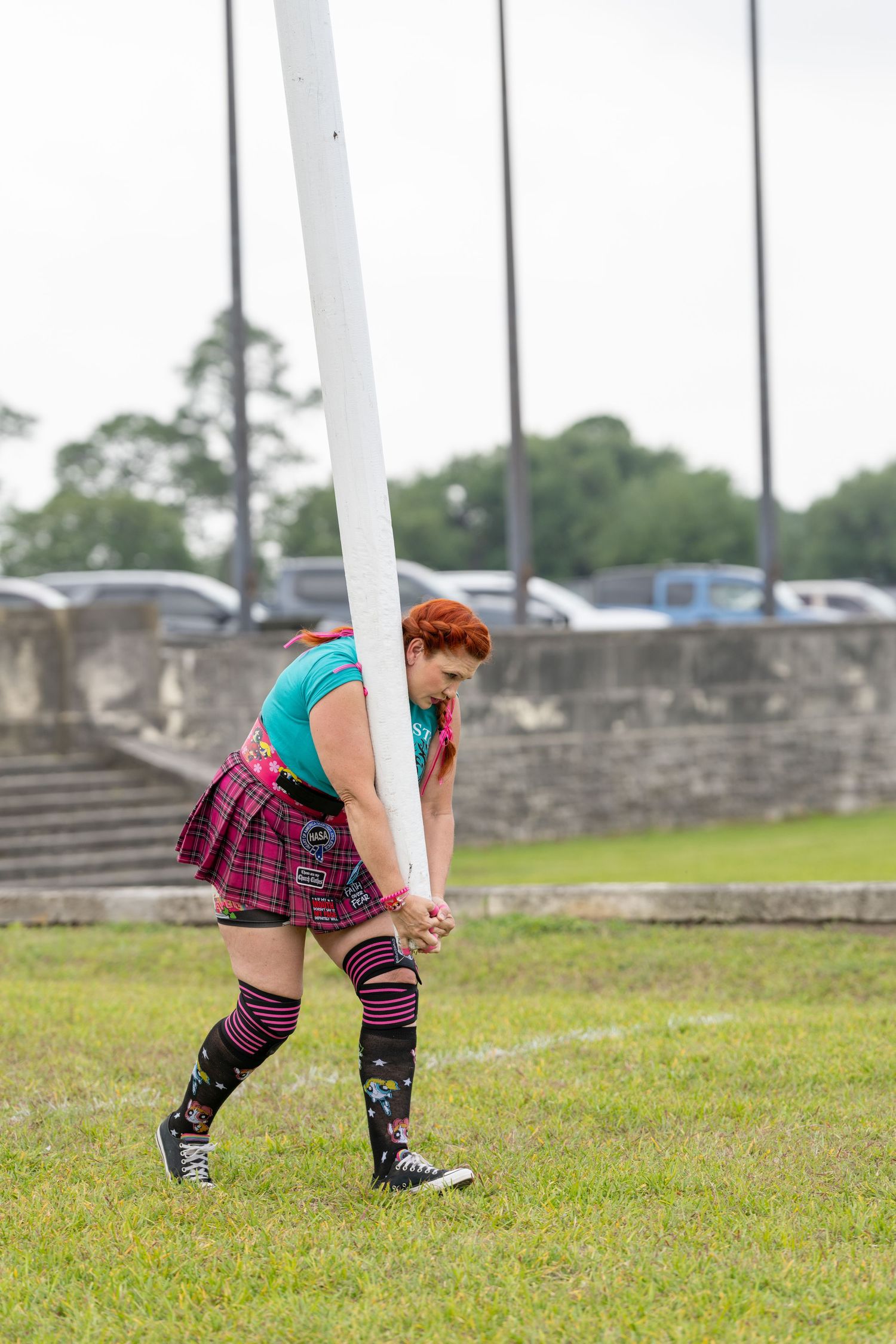 Woman in plaid skirt and colorful socks tossing a caber at a Highland Games competition outdoors.