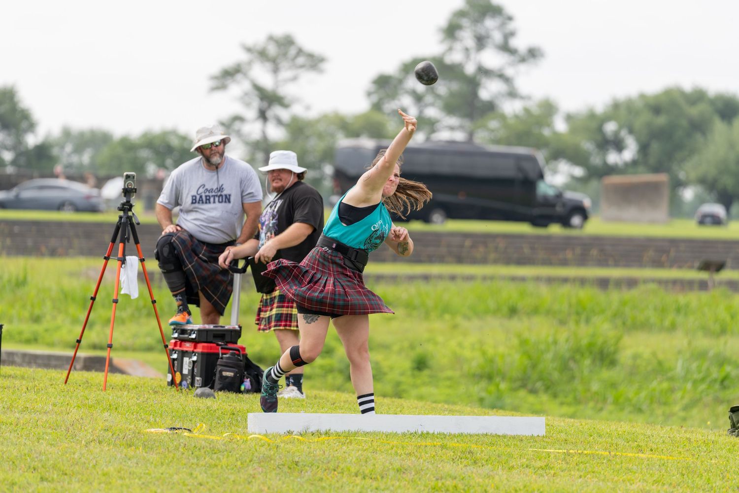 Athlete in tartan kilt competes in Highland Games hammer throw event outdoors, with officials and equipment visible in background.