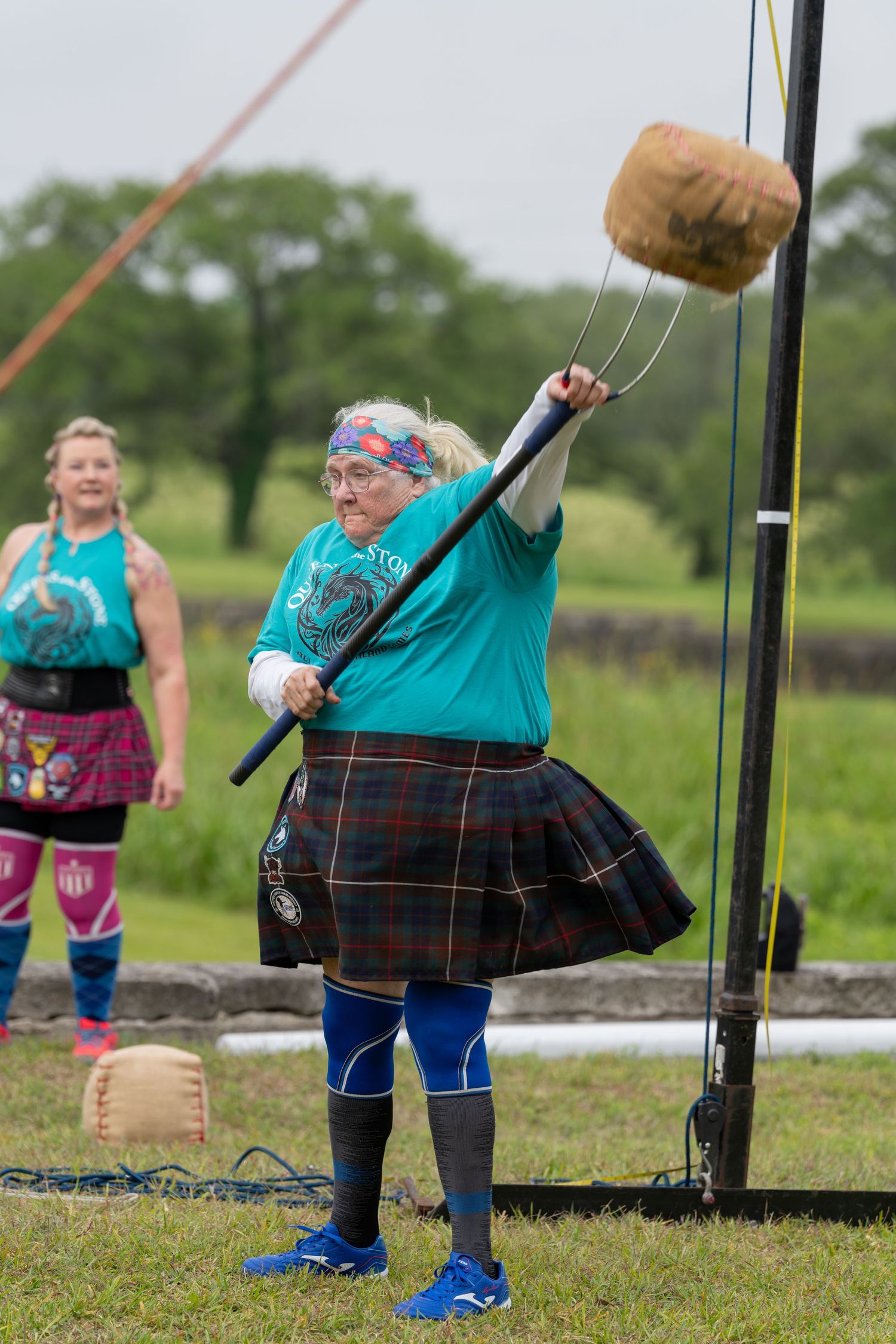 Woman in tartan kilt throws weight over bar at Scottish Highland Games competition.