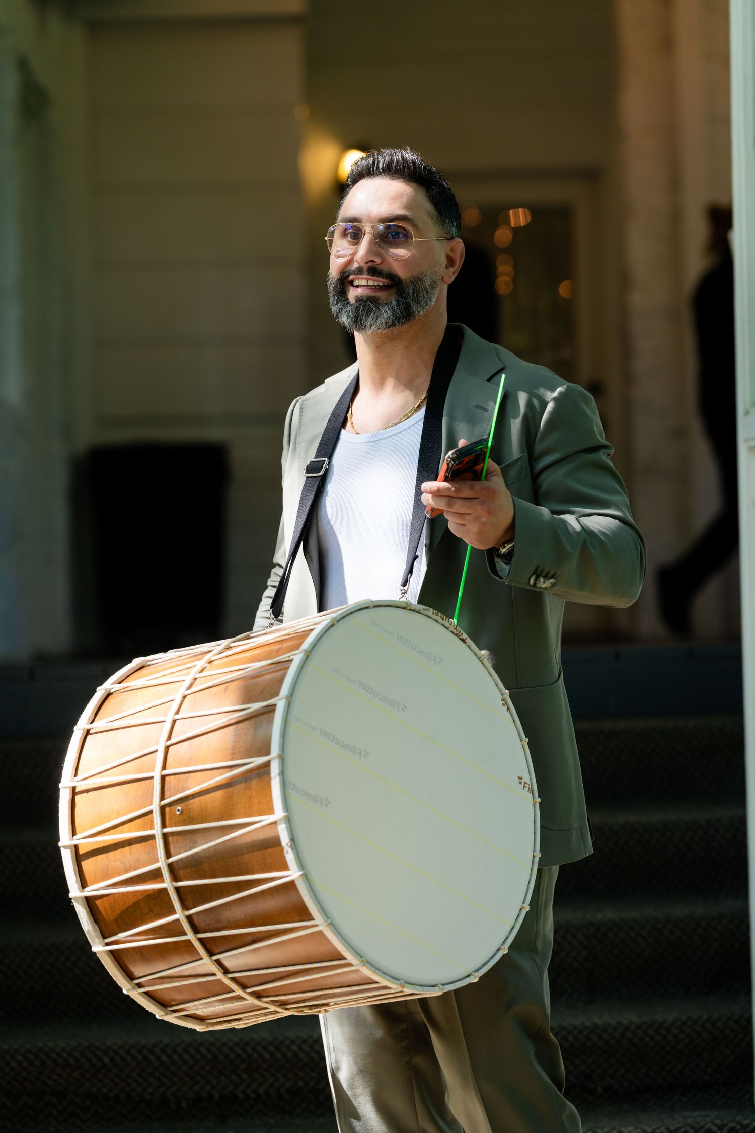 Bearded man in green blazer playing a large drum outdoors on steps, smiling while holding drumsticks.
