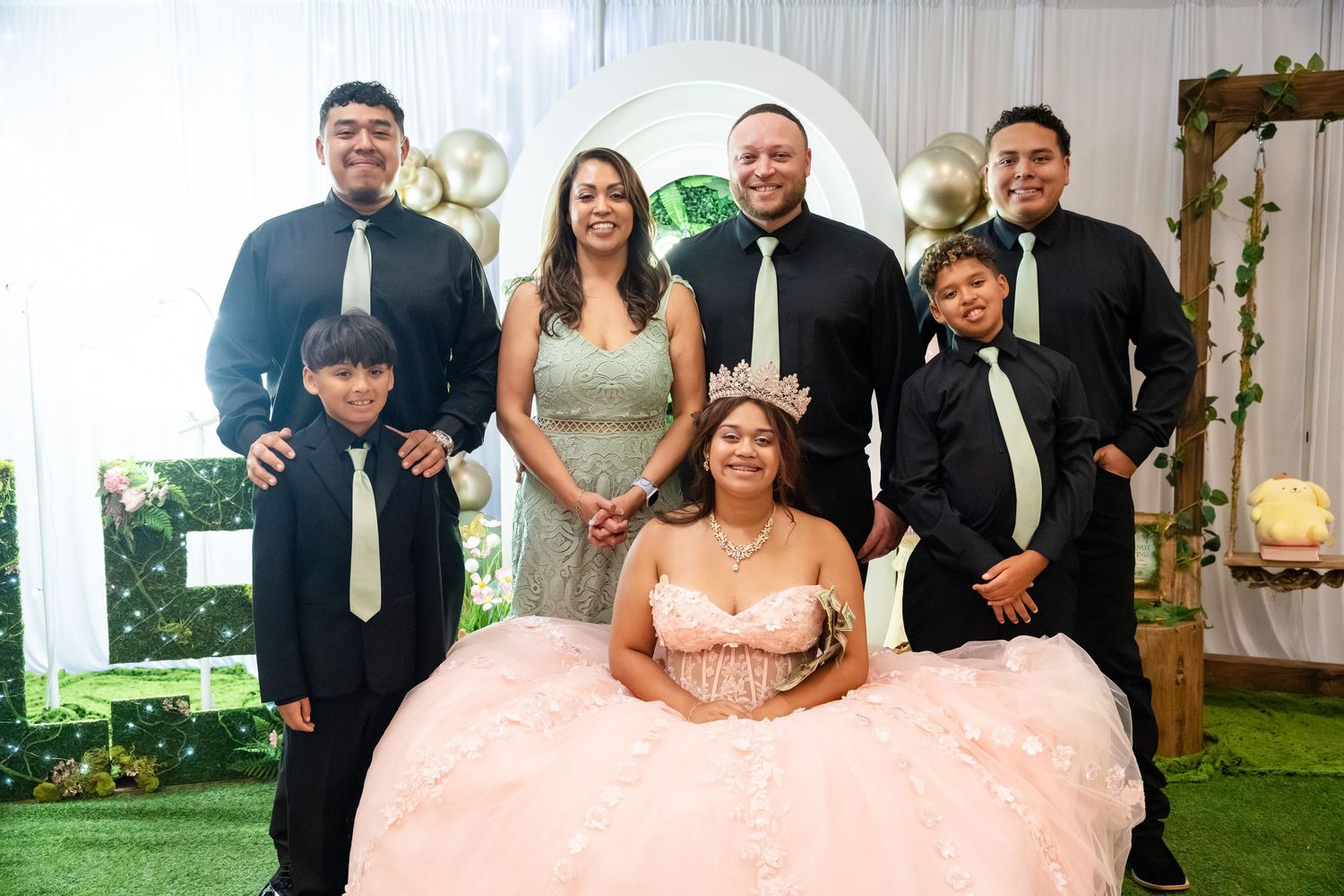 A quinceañera girl in a pink ball gown poses with six family members in formal attire at an elegant decorated venue.