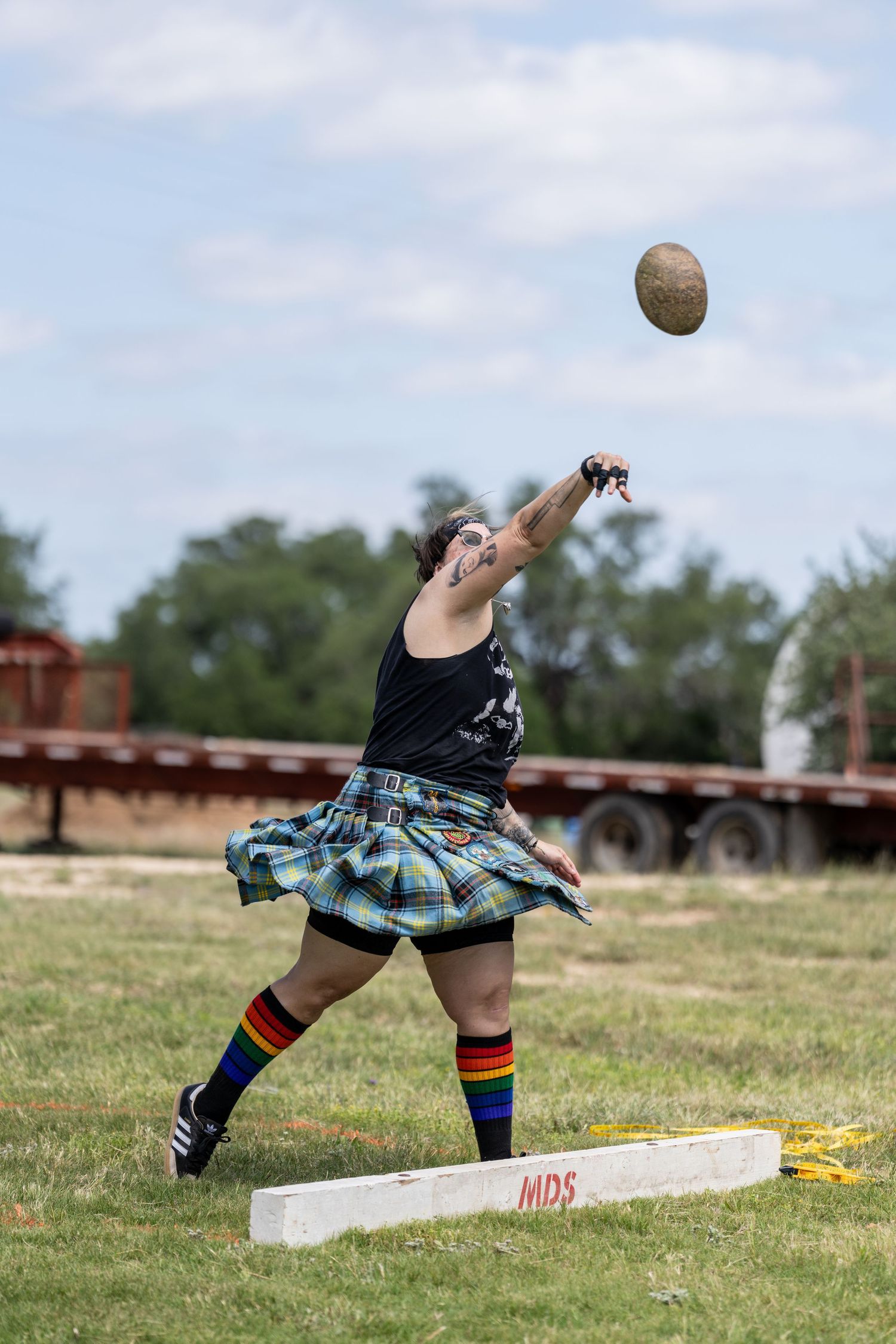 Athlete in tartan kilt throwing stone ball at Highland Games competition outdoors.