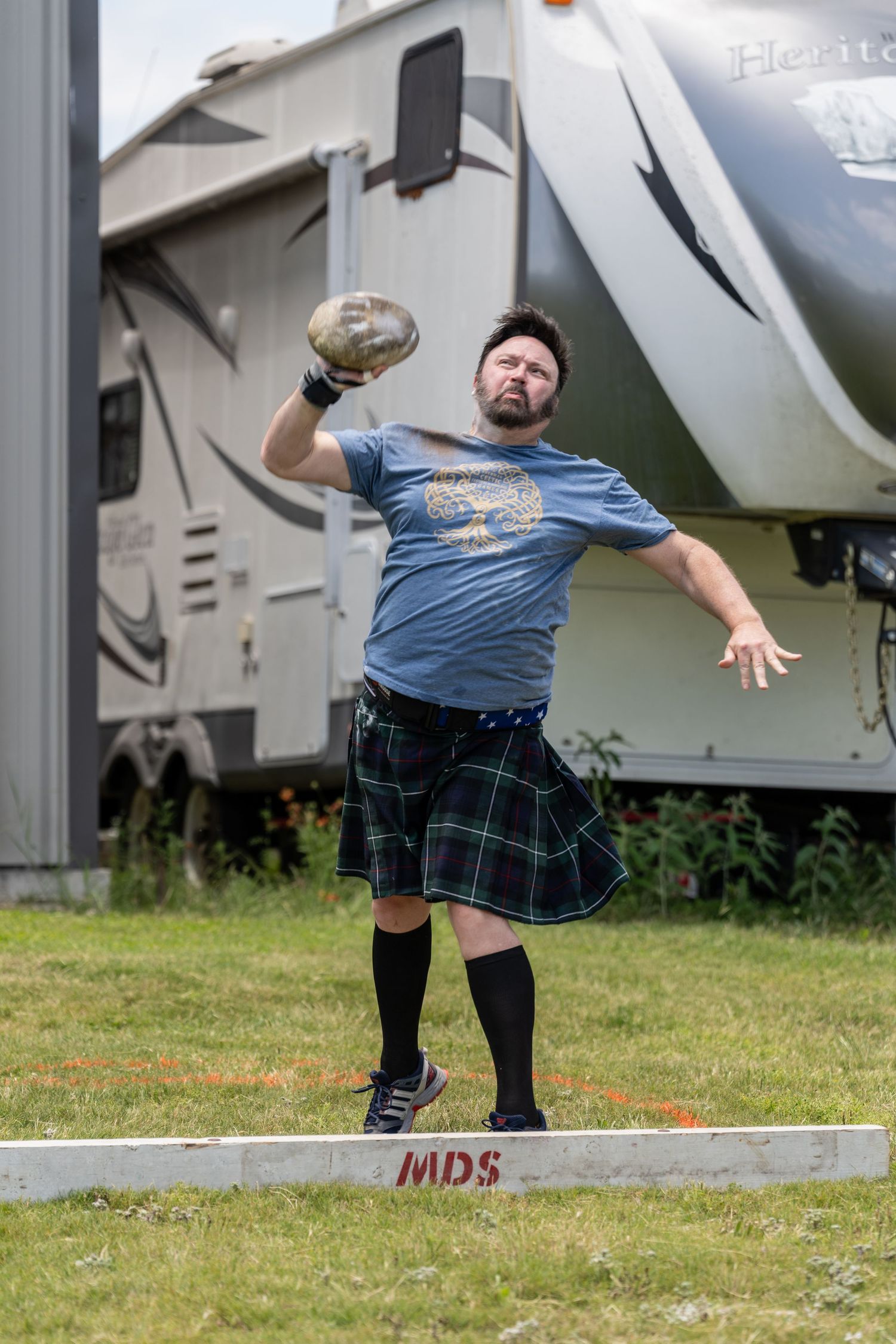 Man in kilt and blue shirt throws shot put at Scottish Highland Games competition outdoors.