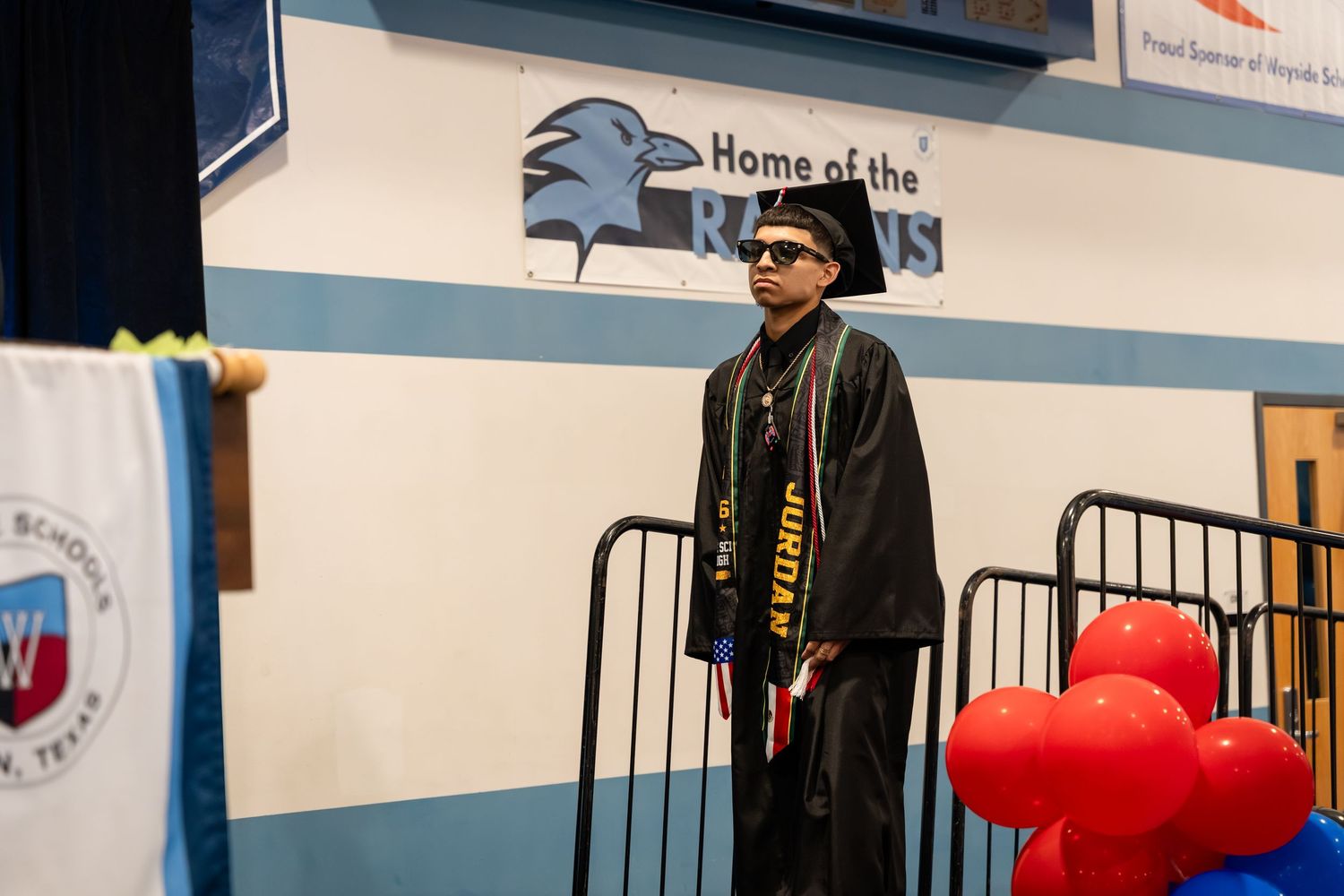 Graduate in cap and gown stands on stage at Ravens school graduation ceremony with balloons.