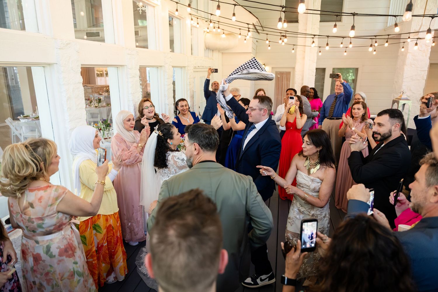 Guests celebrate joyfully at an indoor wedding reception with string lights, dancing and clapping together.