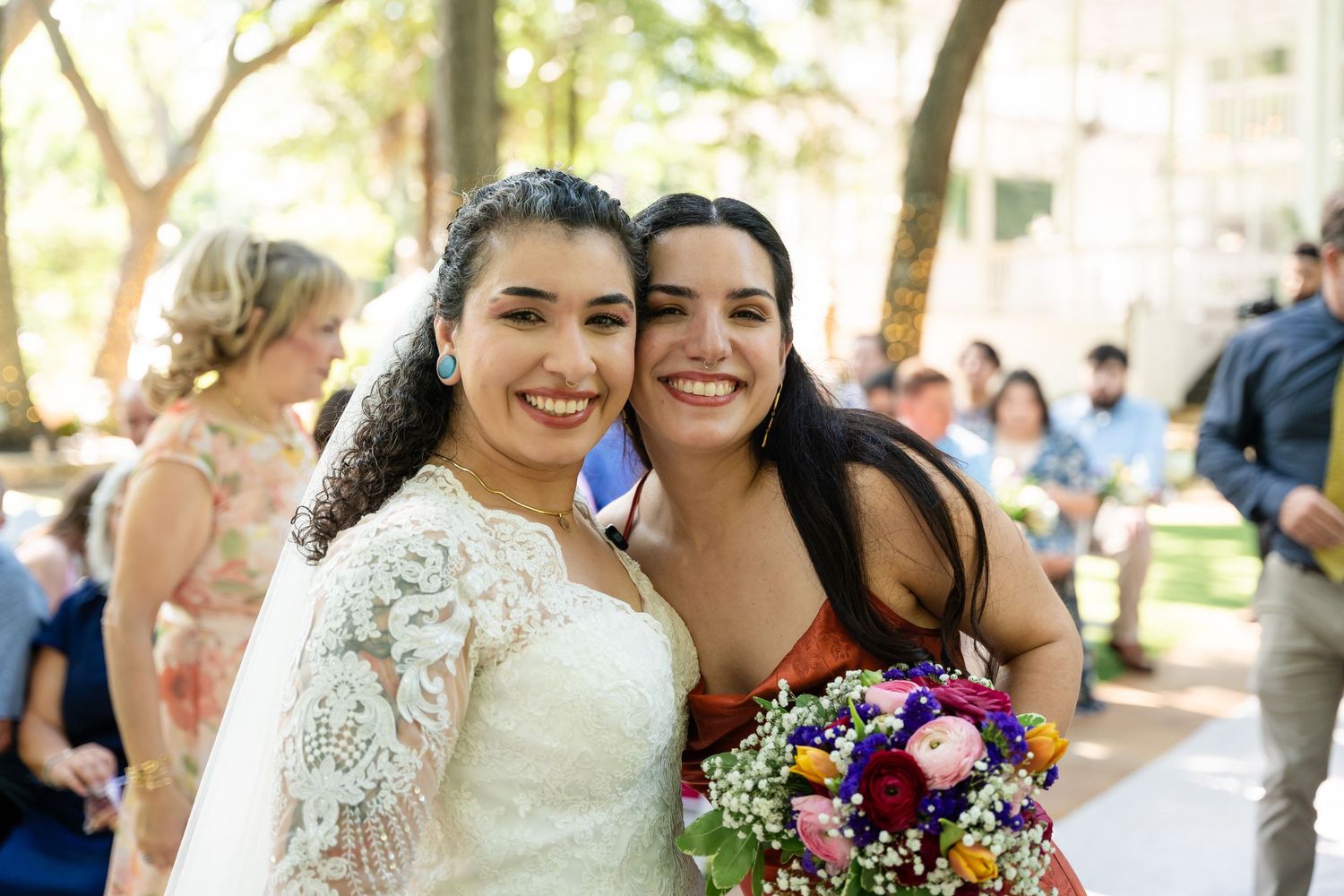 Bride in white lace dress smiles with bridesmaid holding colorful bouquet at outdoor wedding ceremony.