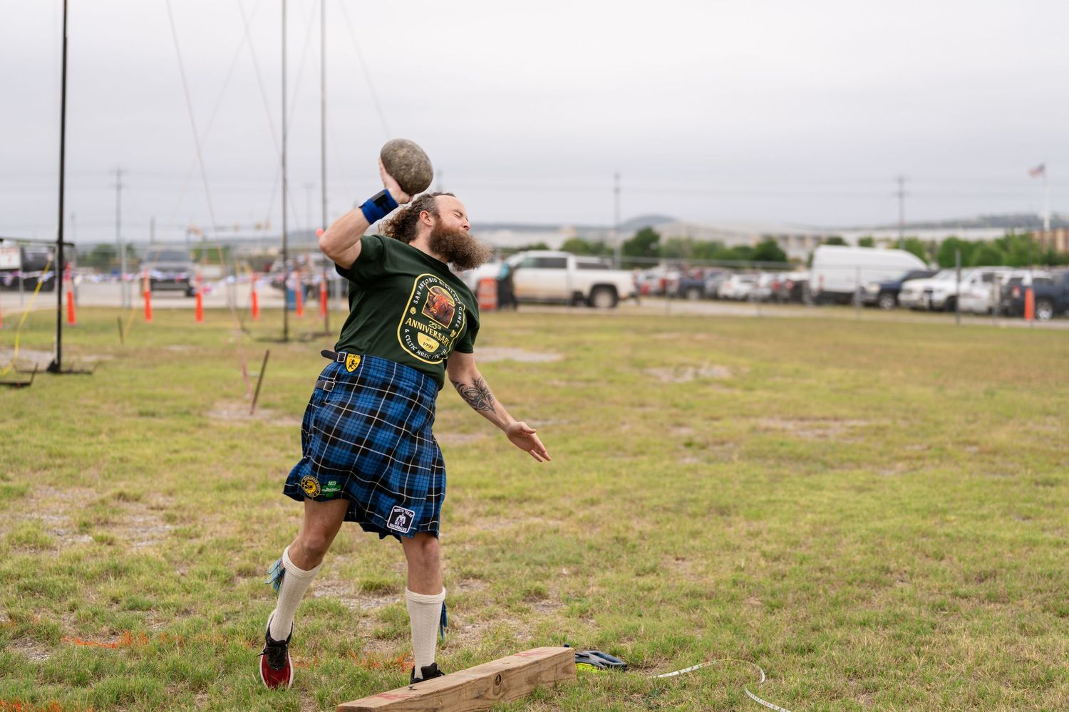 Man in blue tartan kilt throws shot put at Scottish Highland Games outdoor competition.