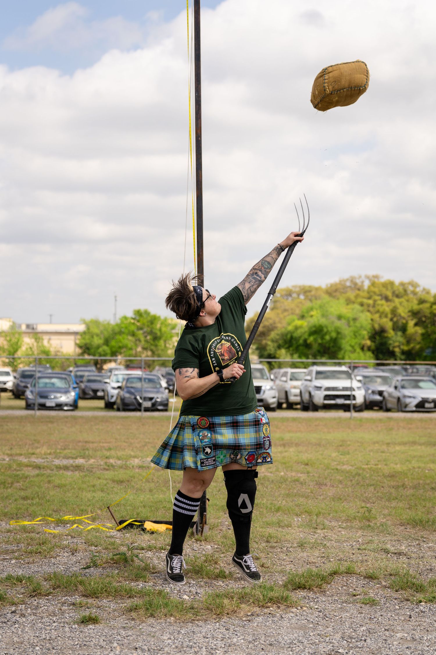 Man in tartan kilt tossing sheaf with pitchfork at Highland Games competition outdoors.