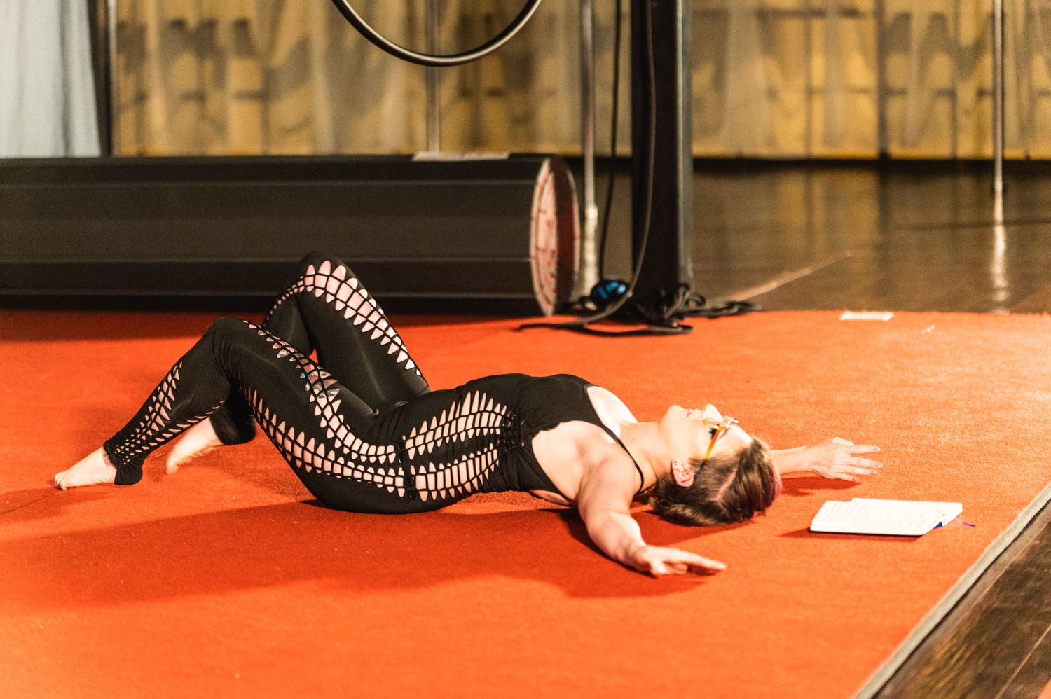 Performer in striped black outfit lying on red floor during aerial dance practice.