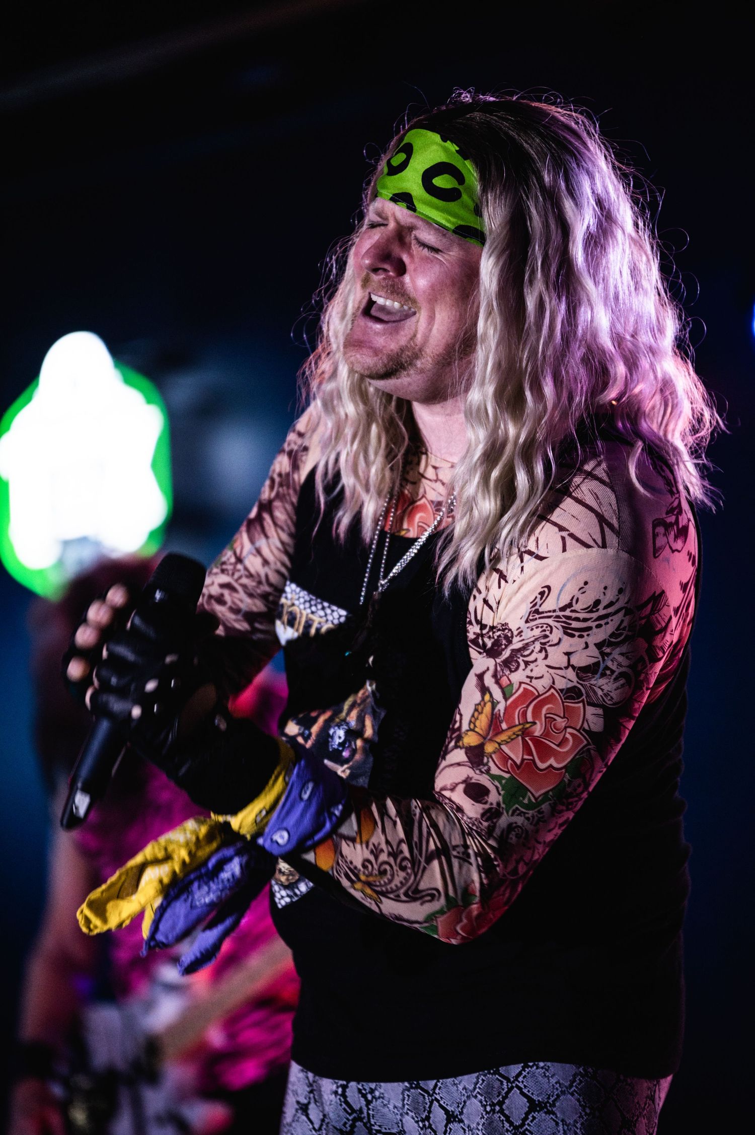 Rock performer with tattooed arms singing passionately under colorful stage lighting.