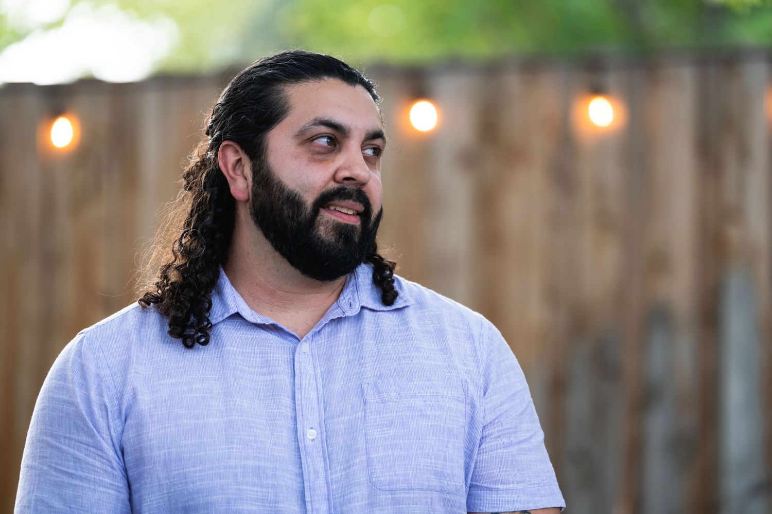 A person with long curly hair and beard wearing a light blue shirt against a backyard fence.