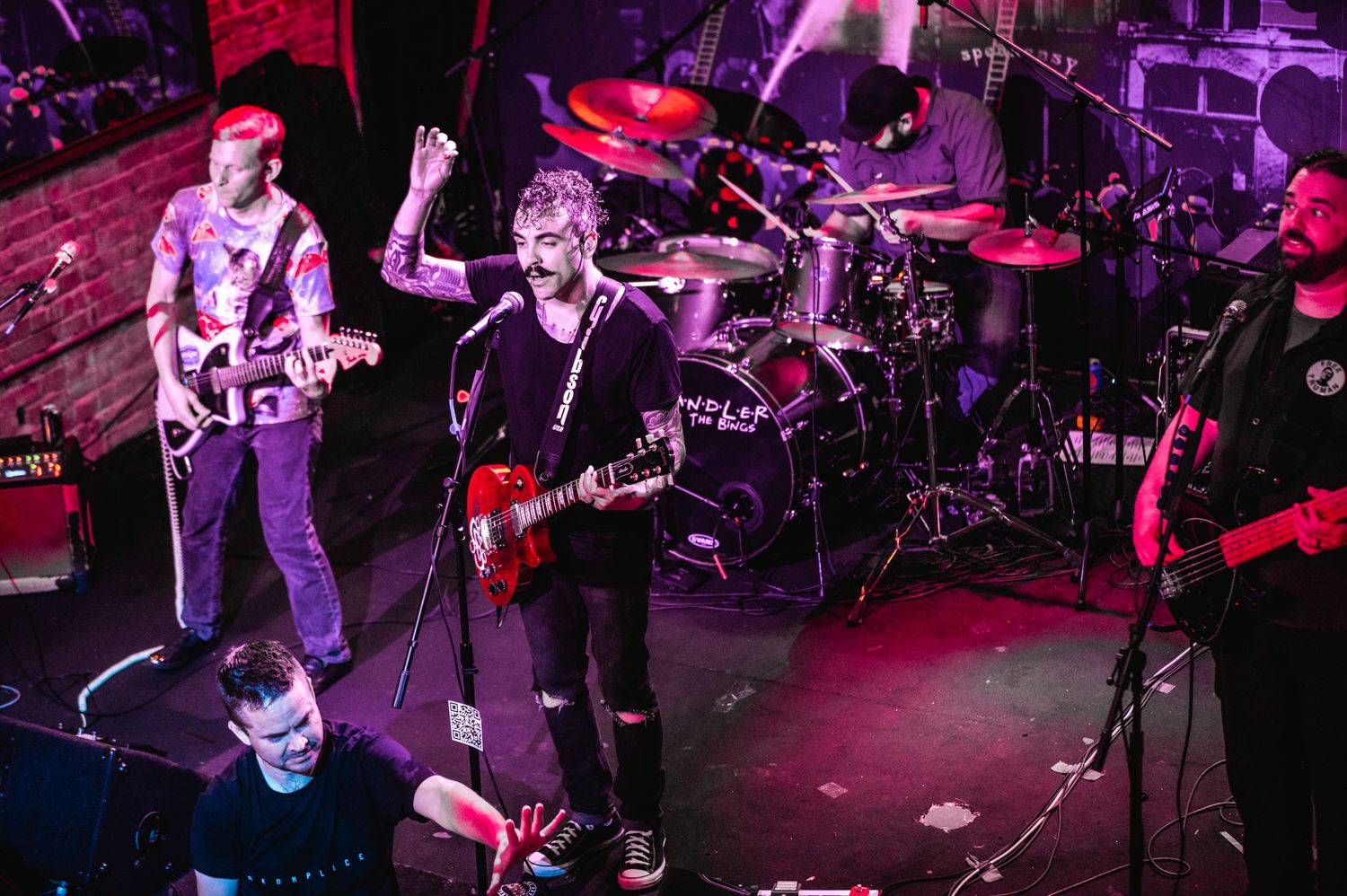 Band performing live with full drum kit and multiple guitarists on dark stage.