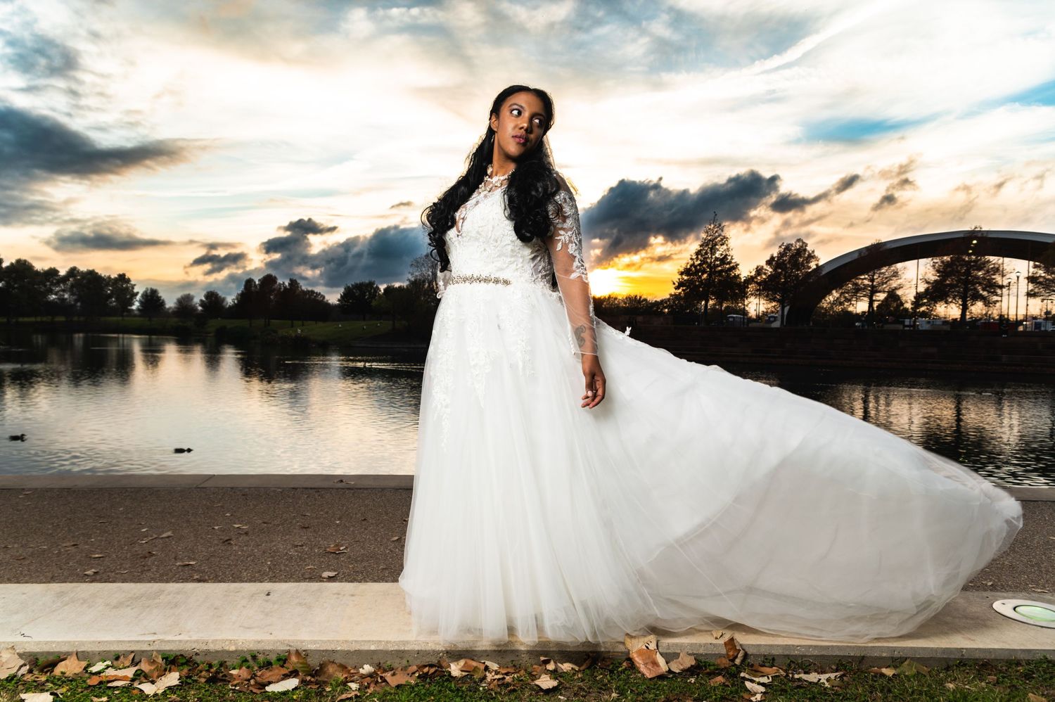 Bride in flowing white gown poses at sunset by lake with dramatic sky.