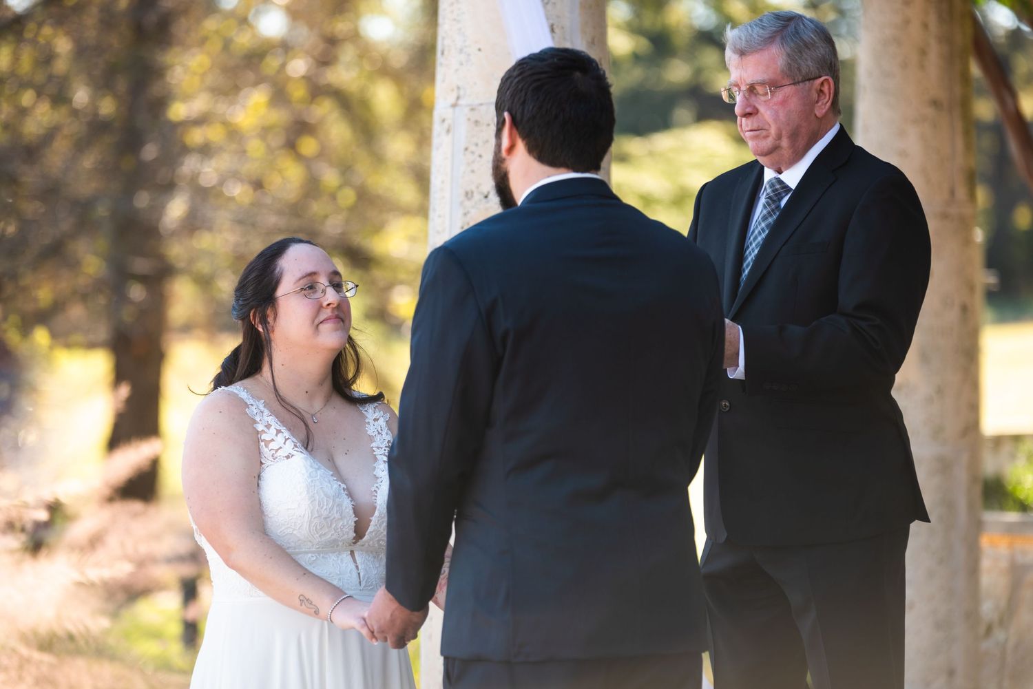 Wedding ceremony under trees with bride in white dress and officiant performing the service.