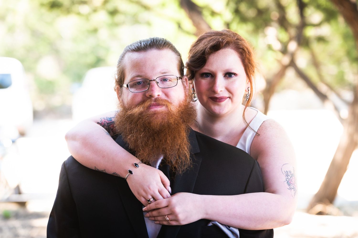 Casual portrait of couple embracing outdoors with sunlit trees in background.