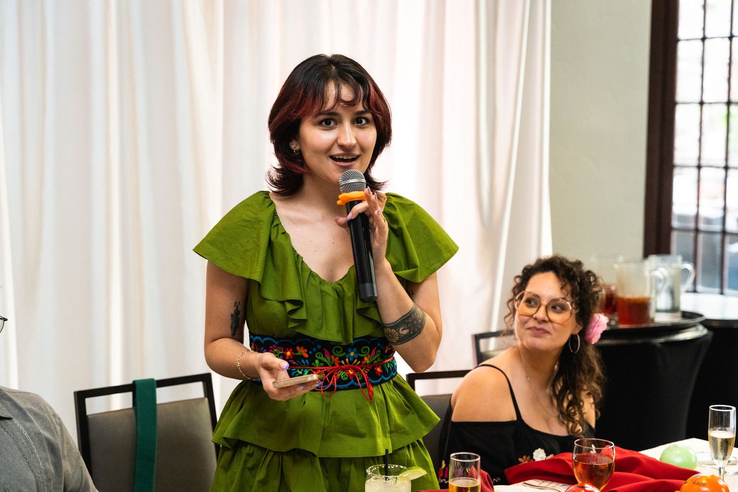 Someone in a green ruffled dress speaks at a celebration event with colorful decorations.