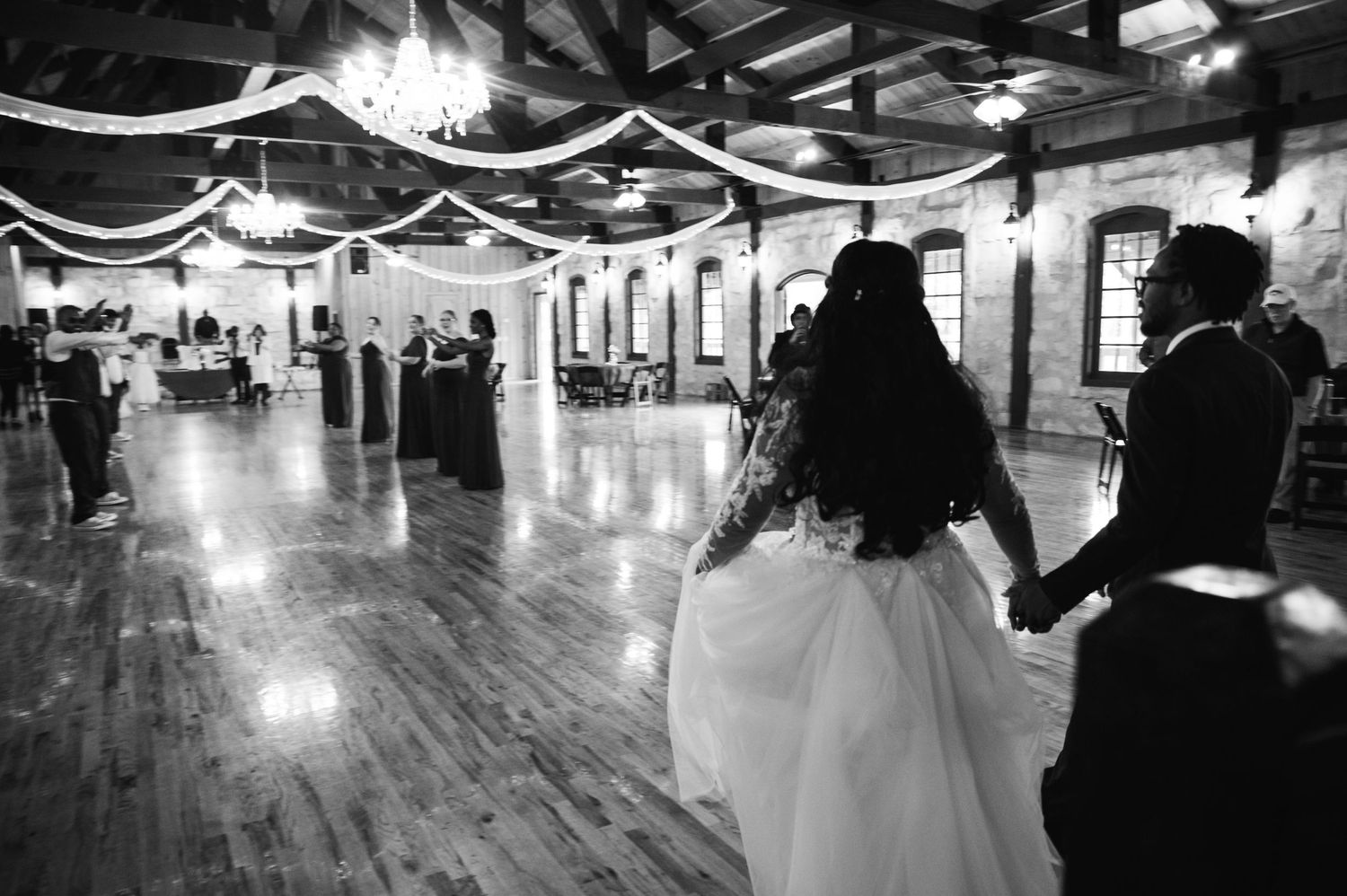 Black and white photo of wedding reception in rustic venue with string lights and wooden floors.