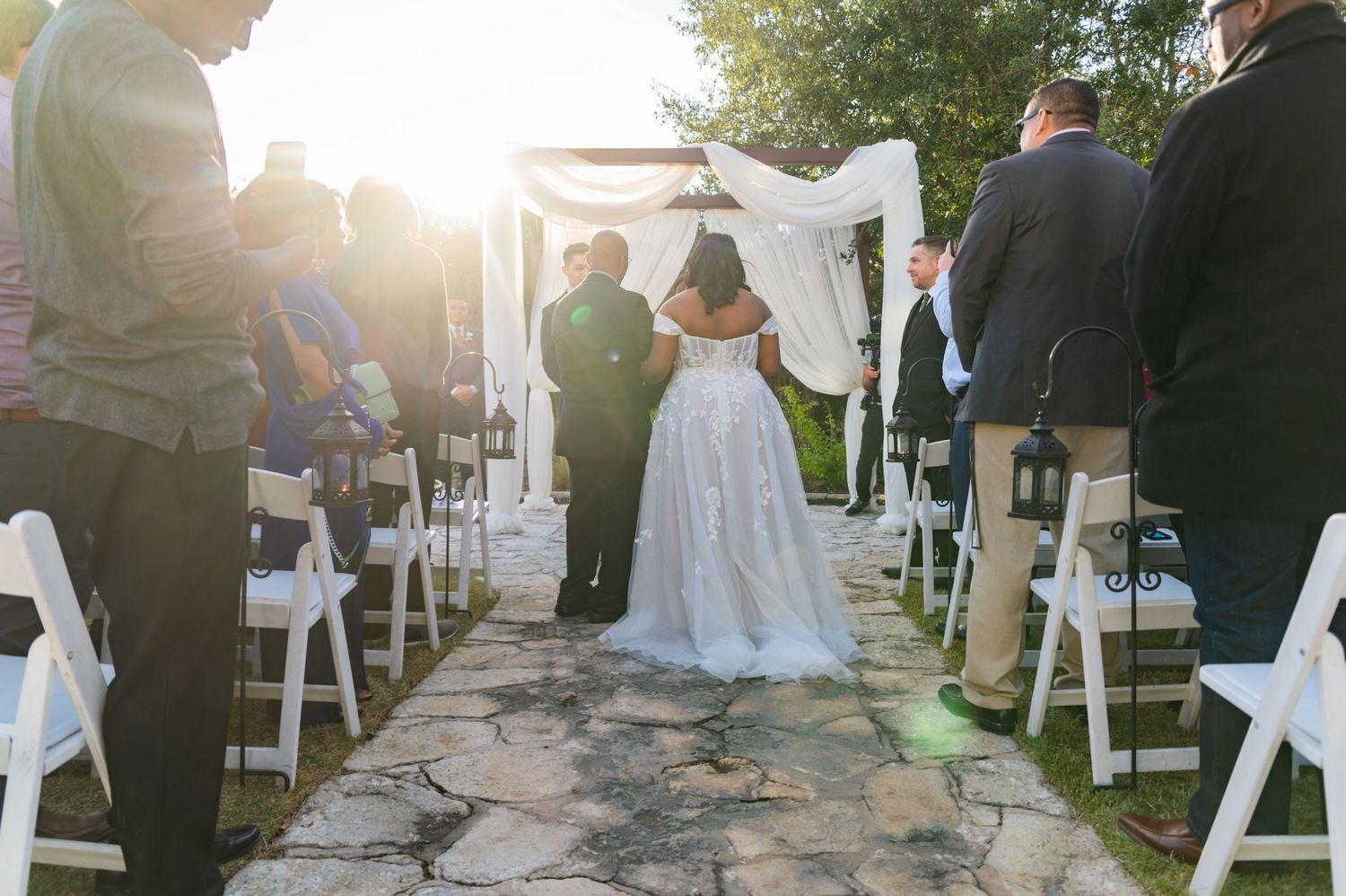 Wedding ceremony in progress at outdoor venue with stone pathway and white chairs.