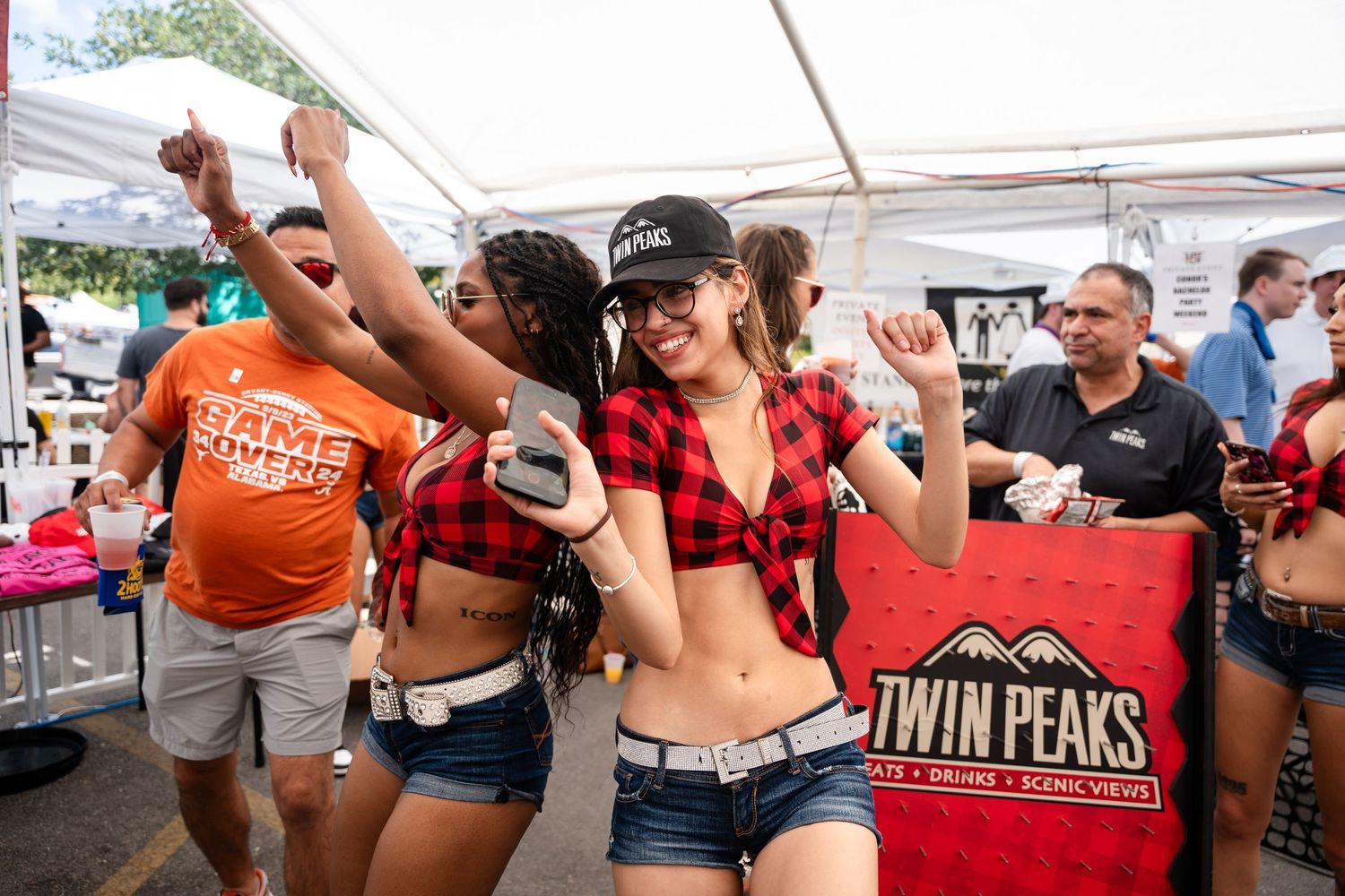 Festive crowd enjoying a lively outdoor event with orange t-shirts and plaid shirts under a white tent.