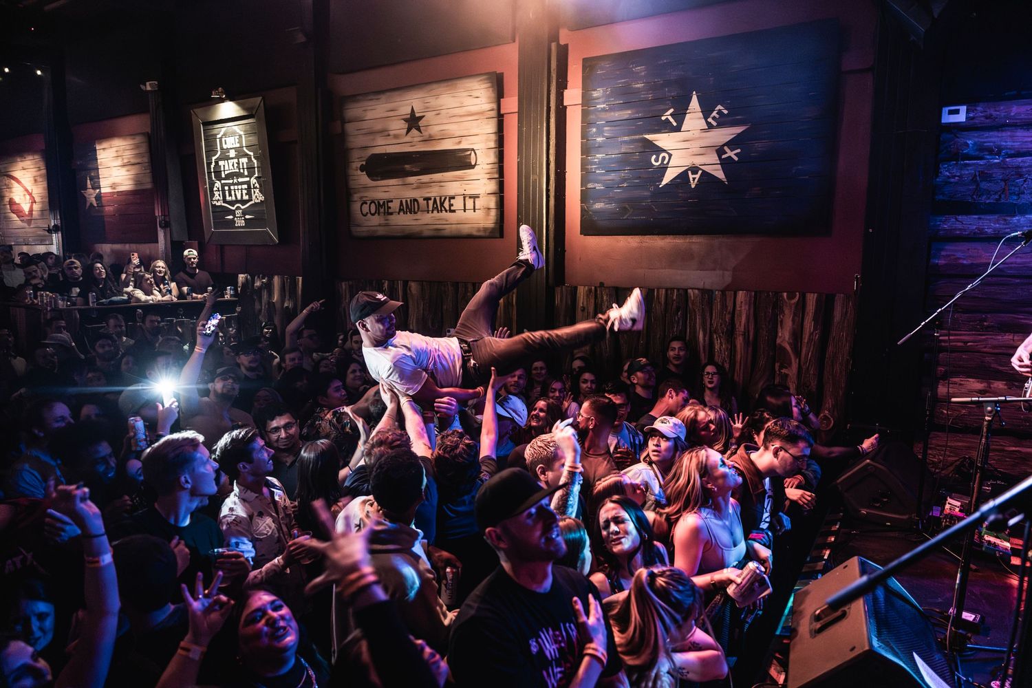 Crowded music venue with Texas flag on wall and energetic audience.