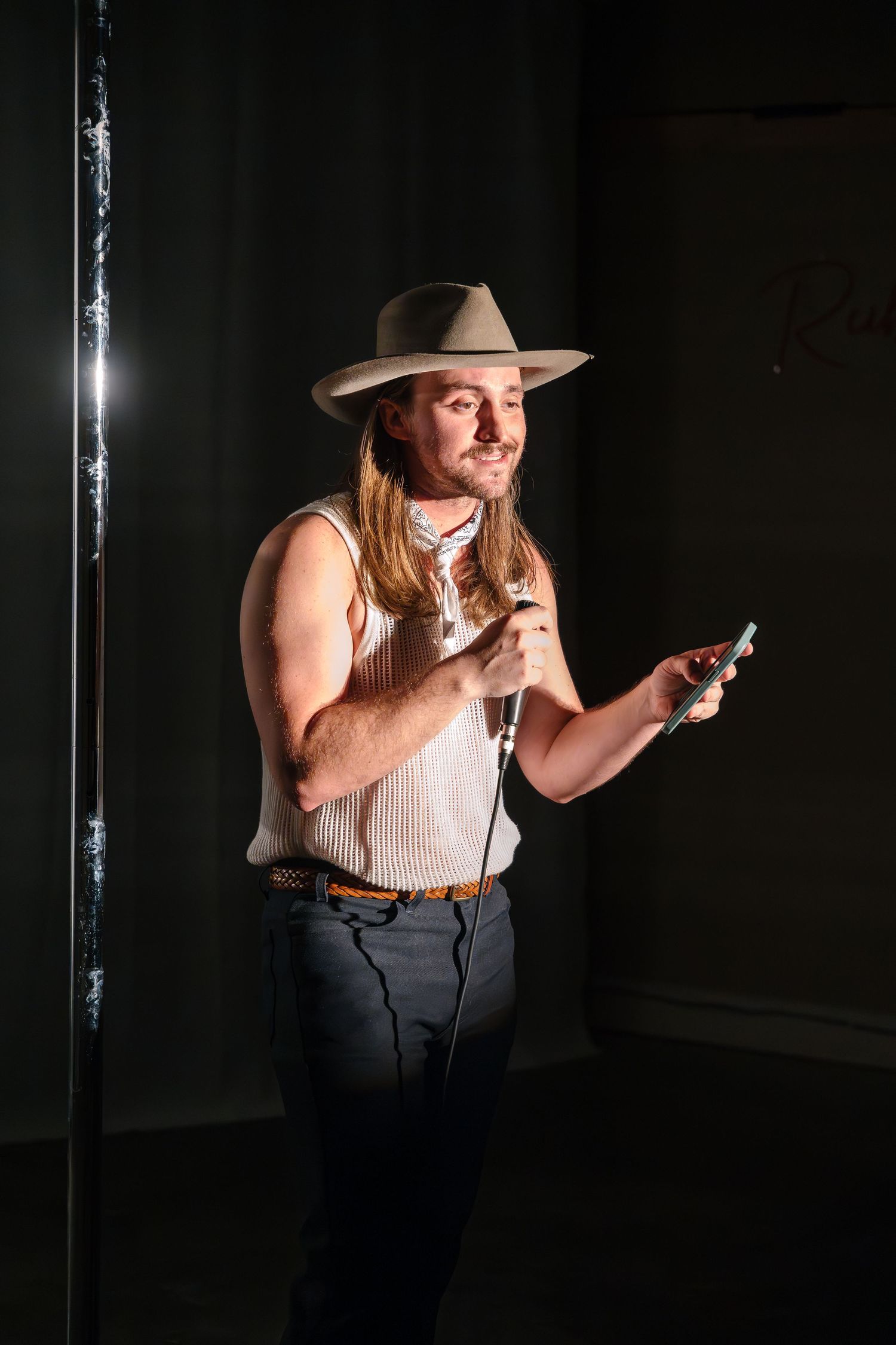 Performer in western attire and hat holds props while standing on a dark stage.