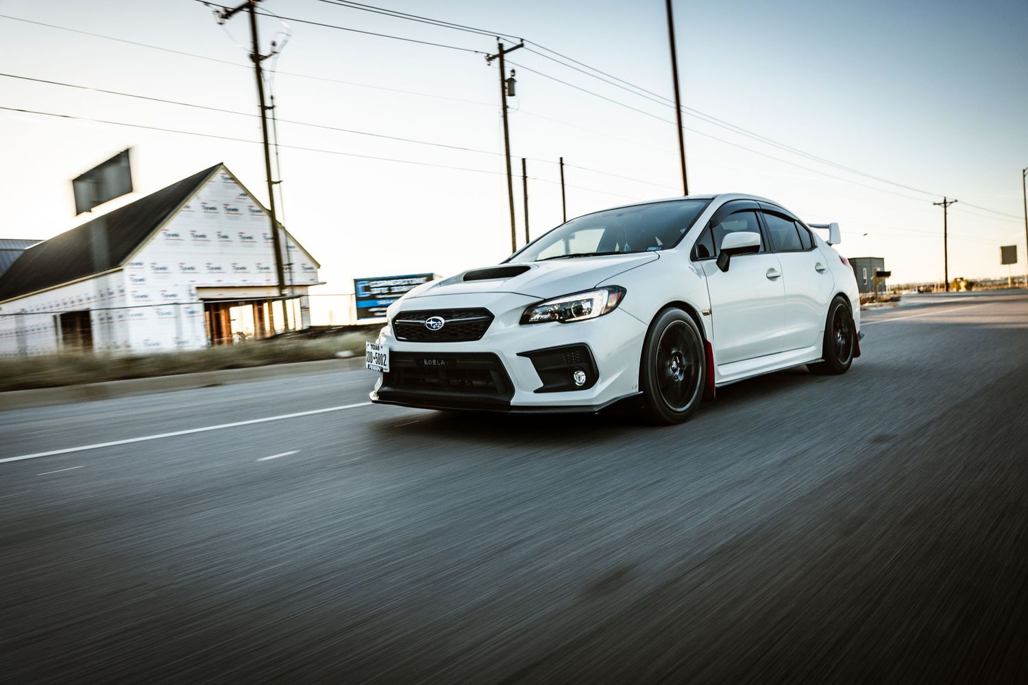 White Subaru WRX STI with aftermarket wheels captured in motion on an empty street.