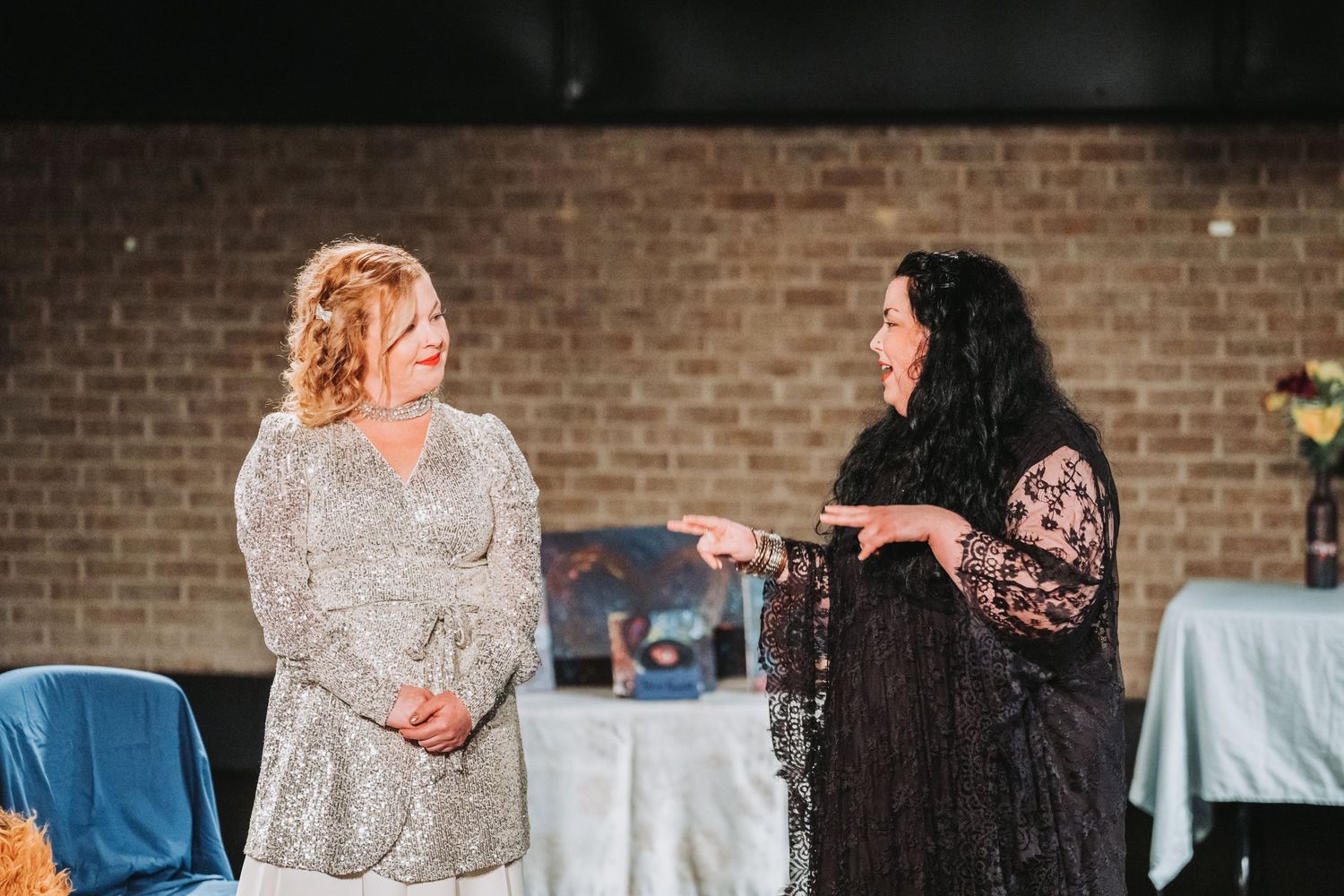 Two performers in elegant dresses share a moment during a theatrical scene.