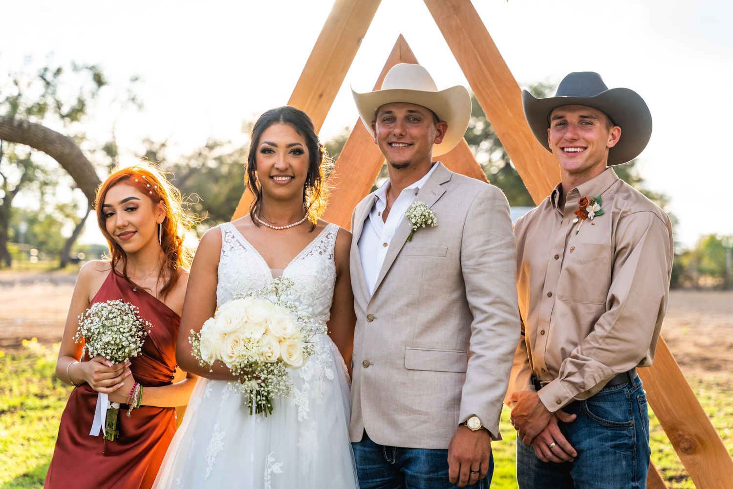 Wedding party in western attire poses under wooden triangular arch during outdoor ceremony at sunset.