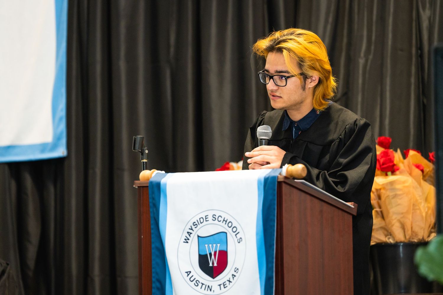 Speaker at podium during academic ceremony with wine glass and yellow hair.
