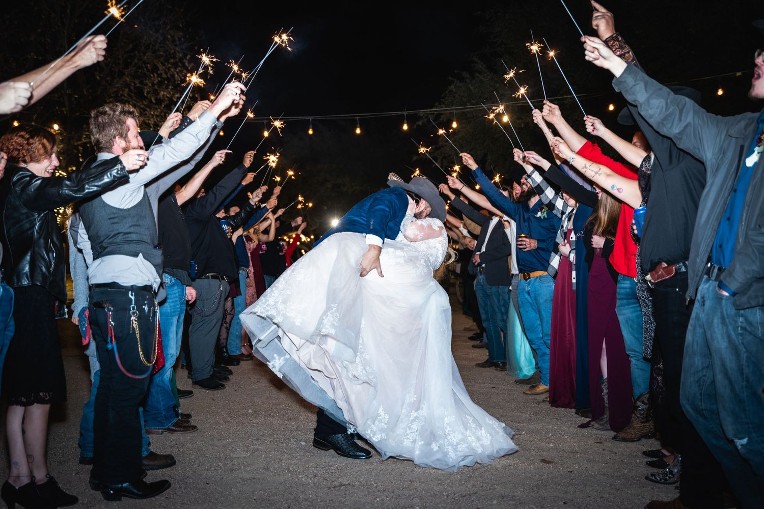 Newlyweds share a romantic dip during their wedding sparkler exit with guests cheering in the background at night.