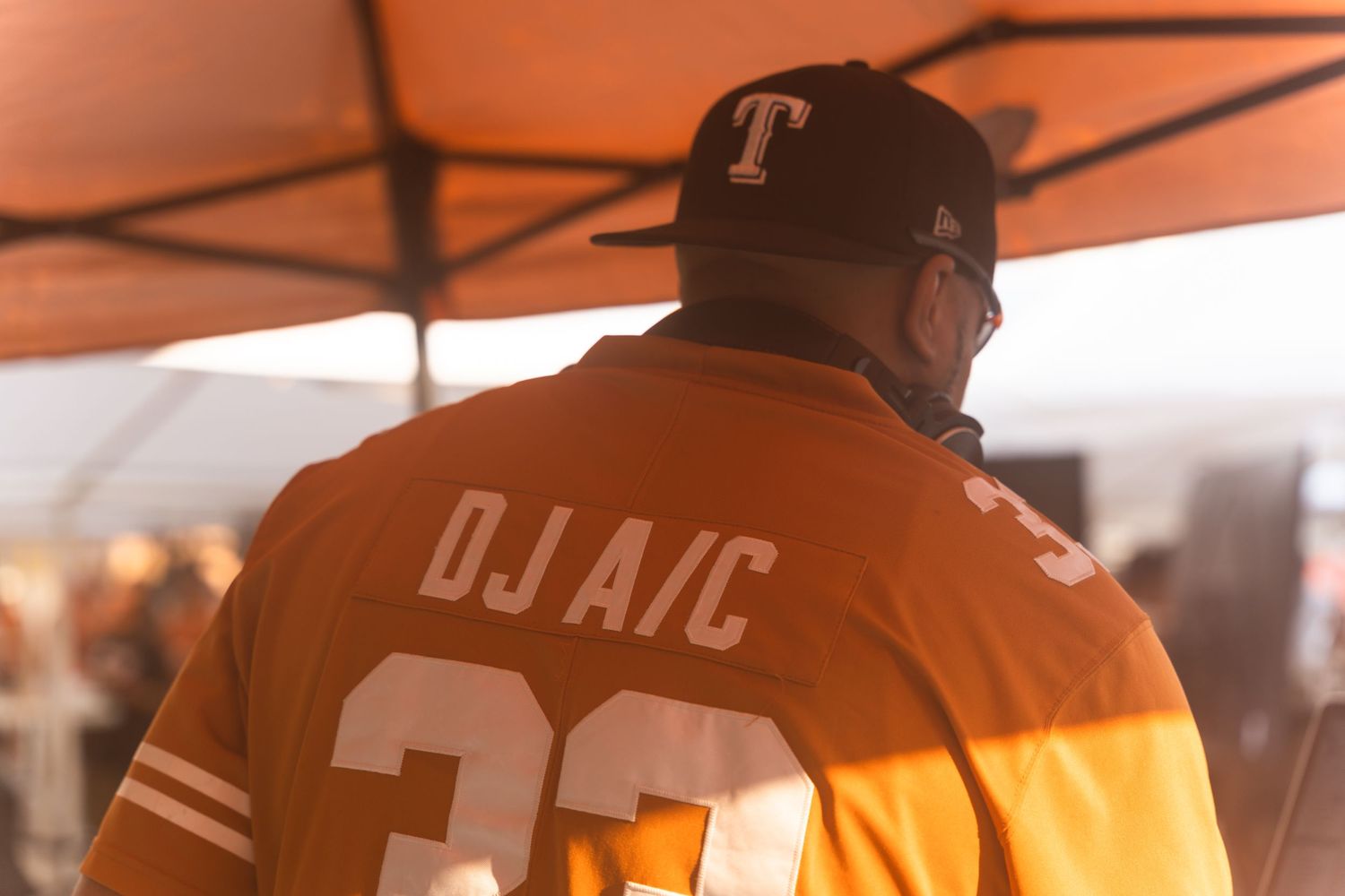 A DJ wearing an orange jersey performs under a tent at an outdoor event.