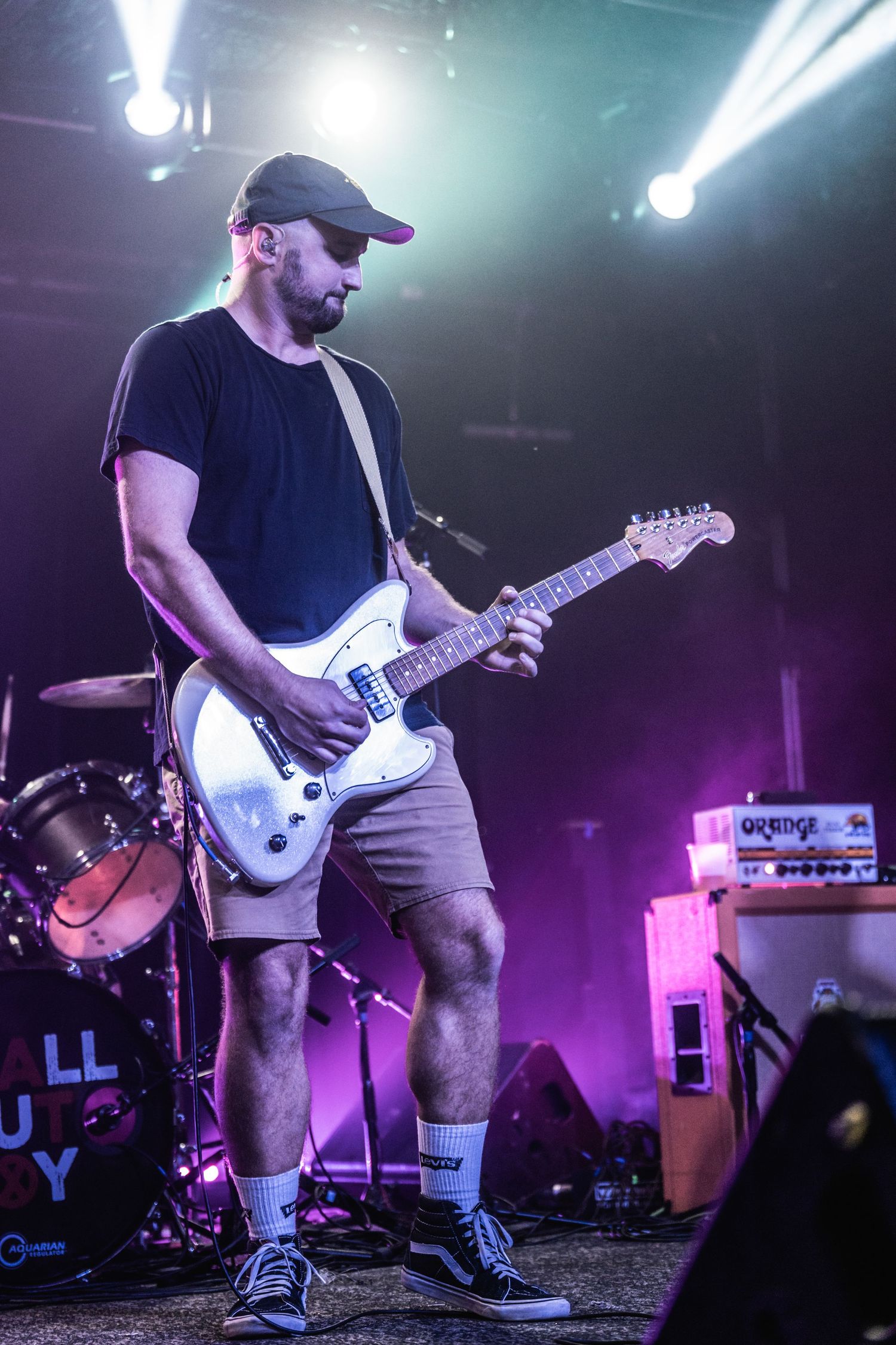 Guitarist performs on stage wearing a black t-shirt and baseball cap under dramatic purple lighting.