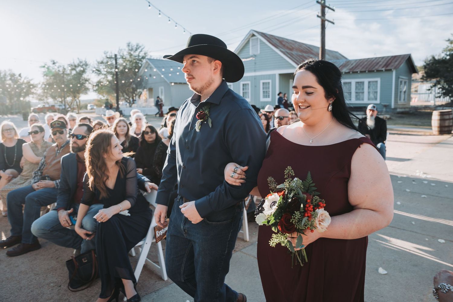 A couple walks together at an outdoor ceremony with guests seated on chairs.